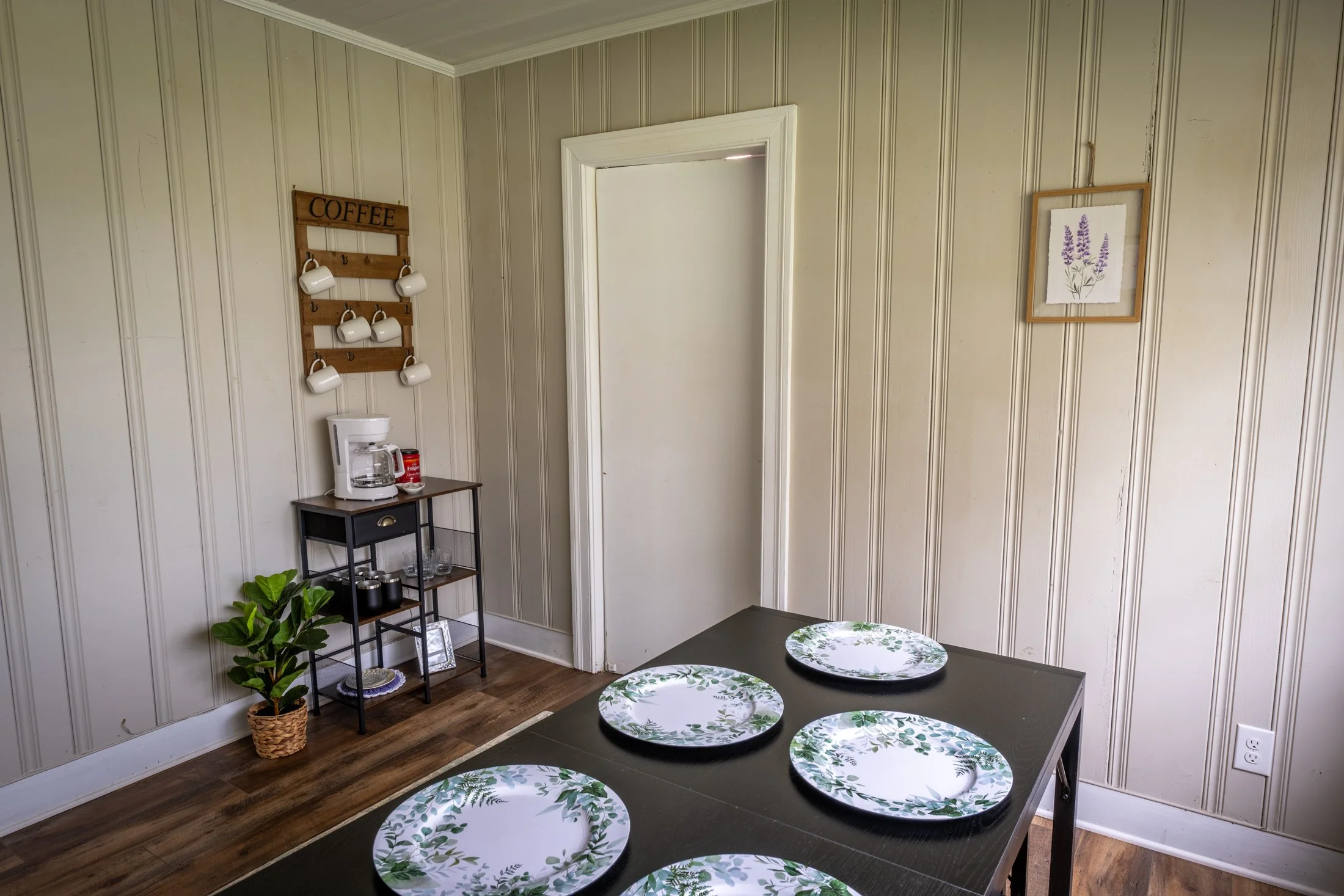 A dining room with a black table set with four white plates with green leaf patterns. On the left, a small corner kitchenette with a coffee maker, a cup rack labeled 'COFFEE,' and a potted plant on the floor. The room has beige vertical wooden wall paneling and hardwood floors.