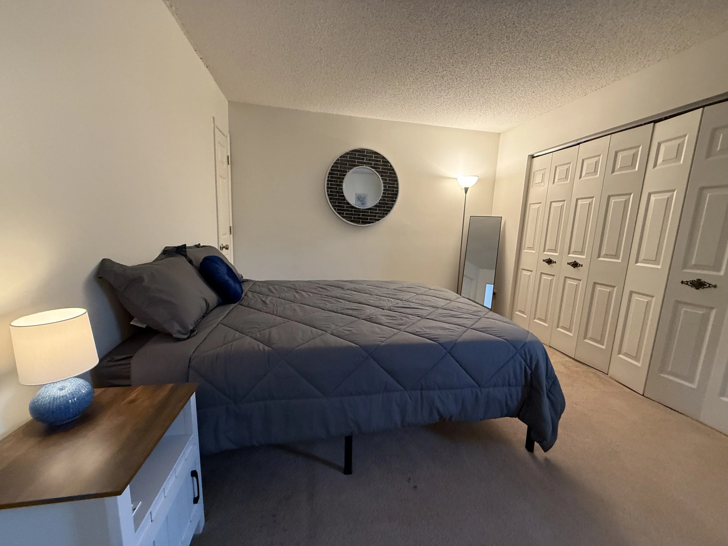 A bedroom with a bed with gray bedding and pillows, a nightstand with a blue vase and a lamp, a round mirror on the wall, a tall floor lamp, and a white closet with double doors.