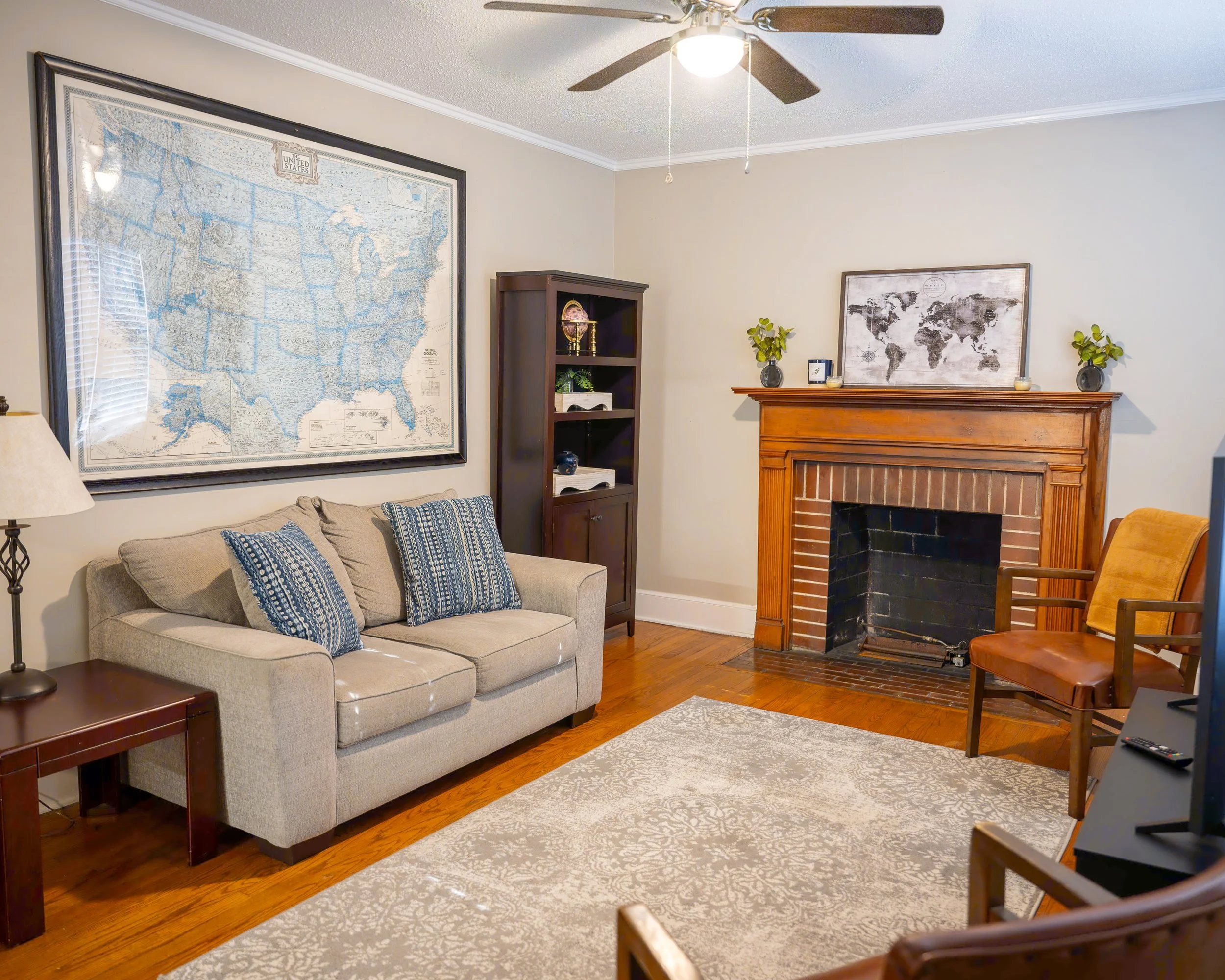 Living room with beige sofa, framed US map, wooden fireplace mantel, brown chair, bookshelf, ceiling fan, and patterned rug.