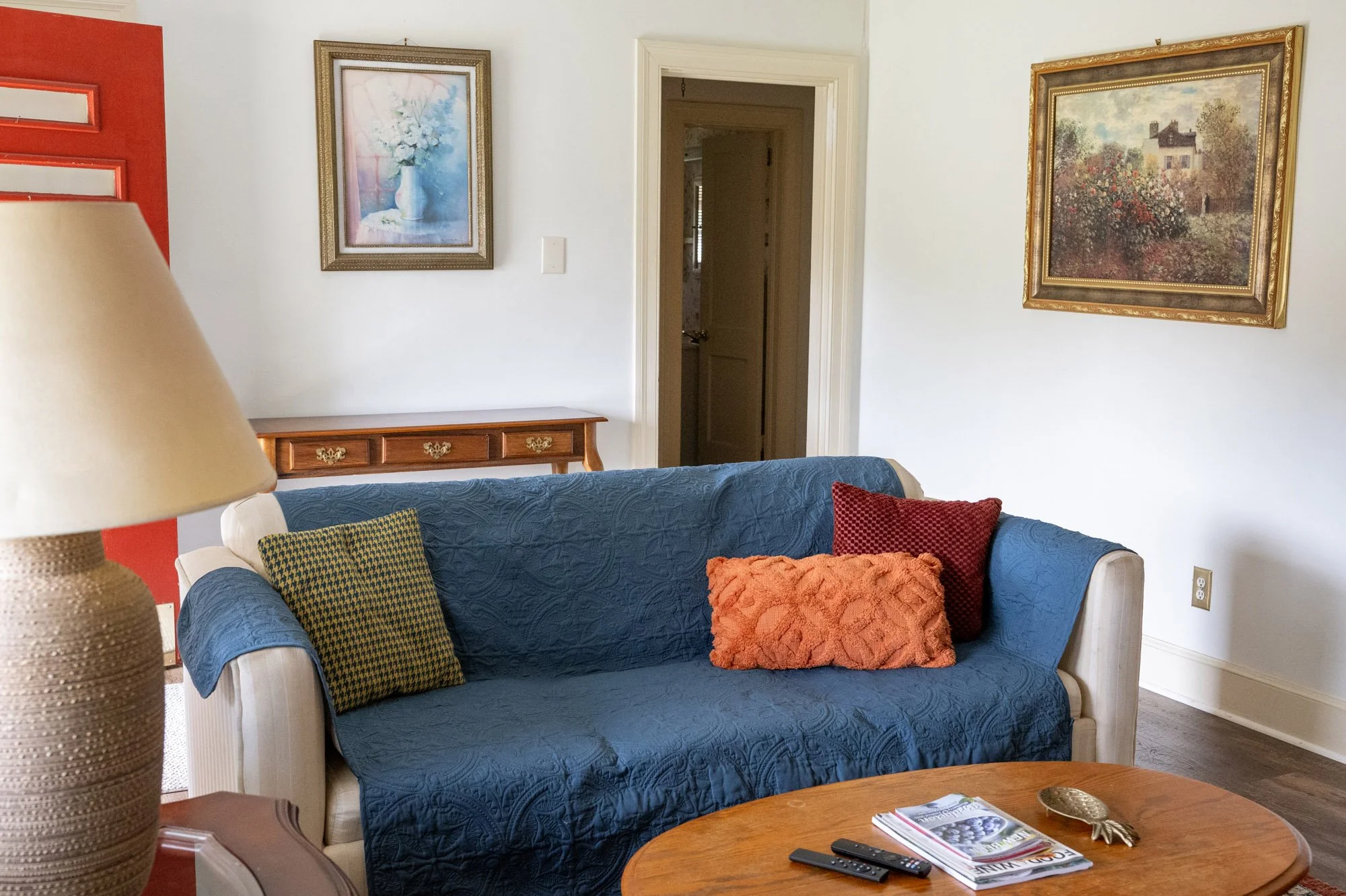 Living room with a beige and blue sofa covered with a blue slipcover, decorated with three throw pillows in red, orange, and yellow-green houndstooth pattern. A wooden coffee table with magazines, remote controls, and a decorative item. A lamp with a beige shade on a side table. Two framed paintings on the wall, one of a white vase with flowers and a landscape scene, and an open doorway leading to another room.
