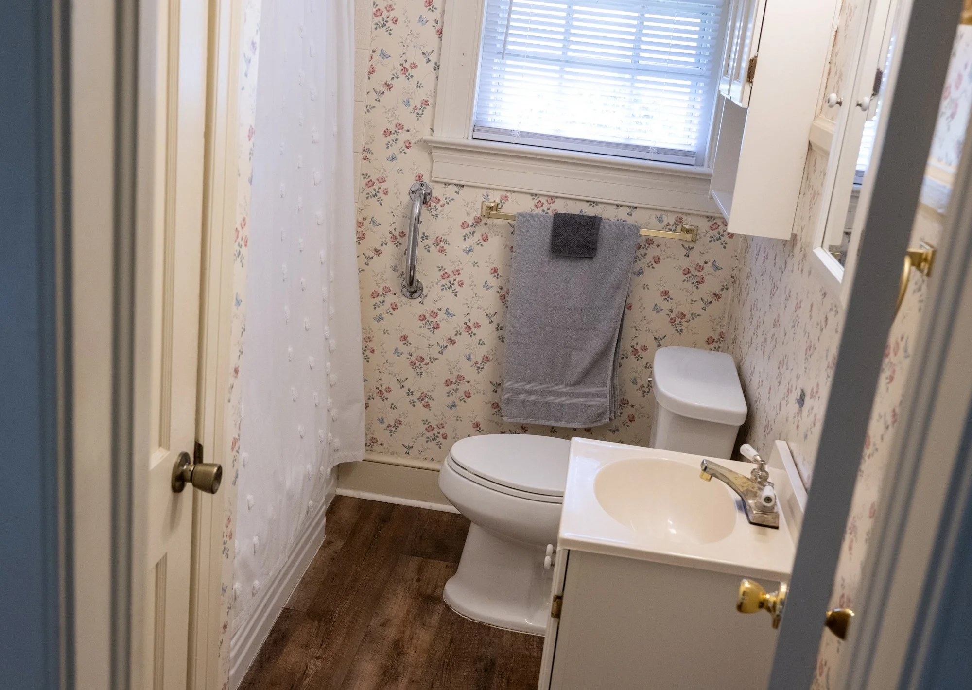 A small bathroom with wood flooring, floral wallpaper, a toilet, a small sink with a gold-cased faucet, a window with blinds, a towel hanging on a bar, and a shower or bathtub with a white curtain.