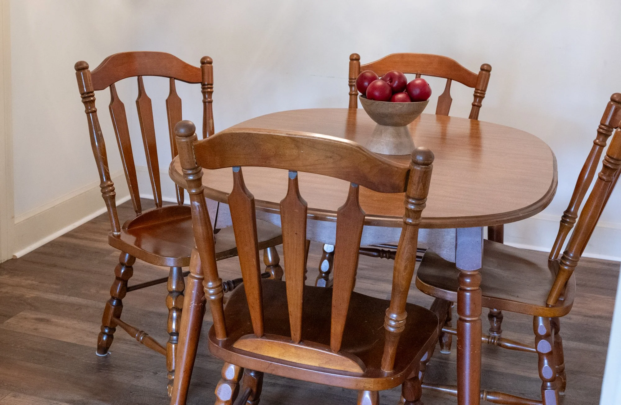 Wooden dining table with five matching wooden chairs, a bowl of red apples on the table, and a hardwood floor with a white wall background.