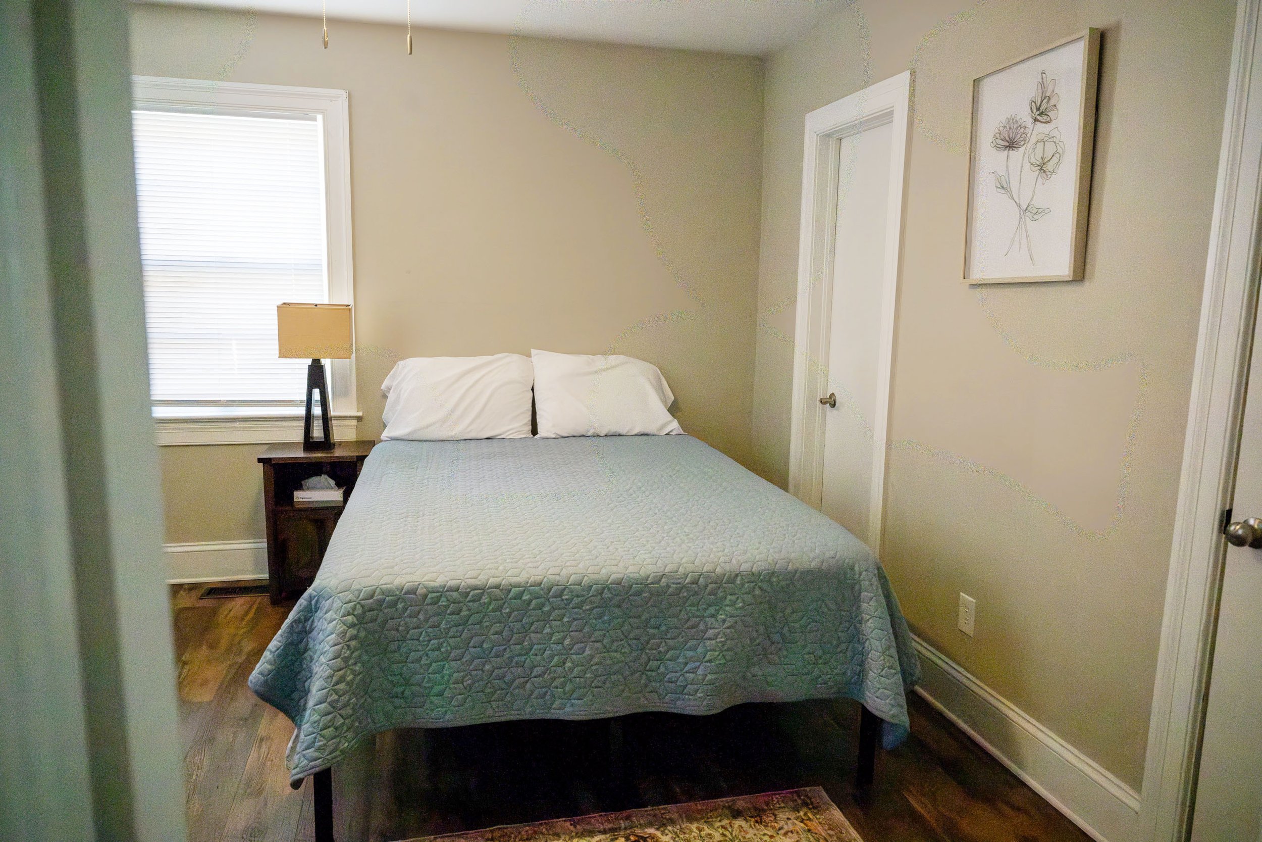 A bedroom with a bed, two white pillows, blue quilt, and a bedside table with a lamp; window with closed blinds and framed floral wall art.
