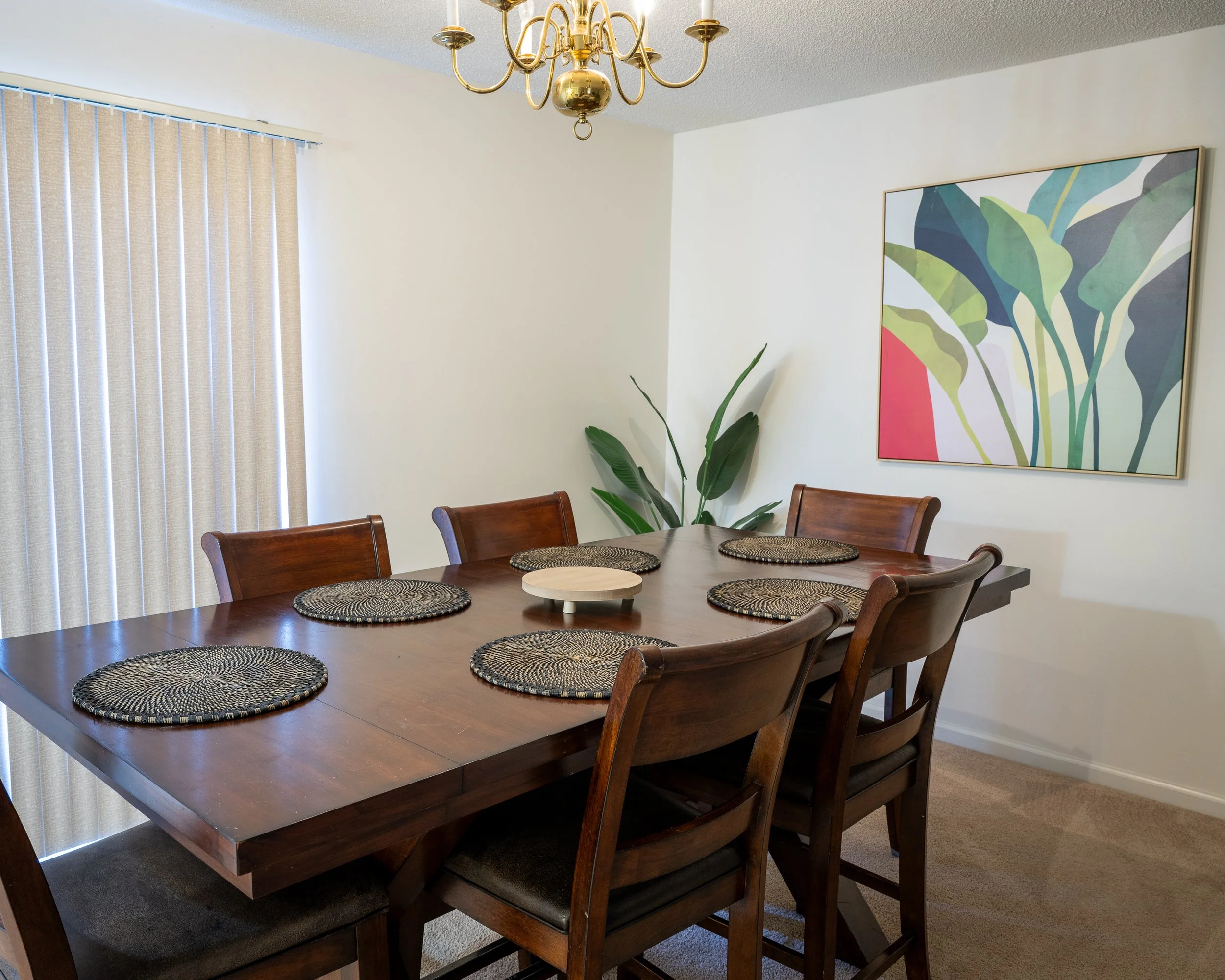 A dining room with a wooden table and six matching chairs, six woven placemats on the table, a potted plant in the corner, a large colorful abstract painting on the wall, beige curtains covering a window, and a brass chandelier hanging above the table.