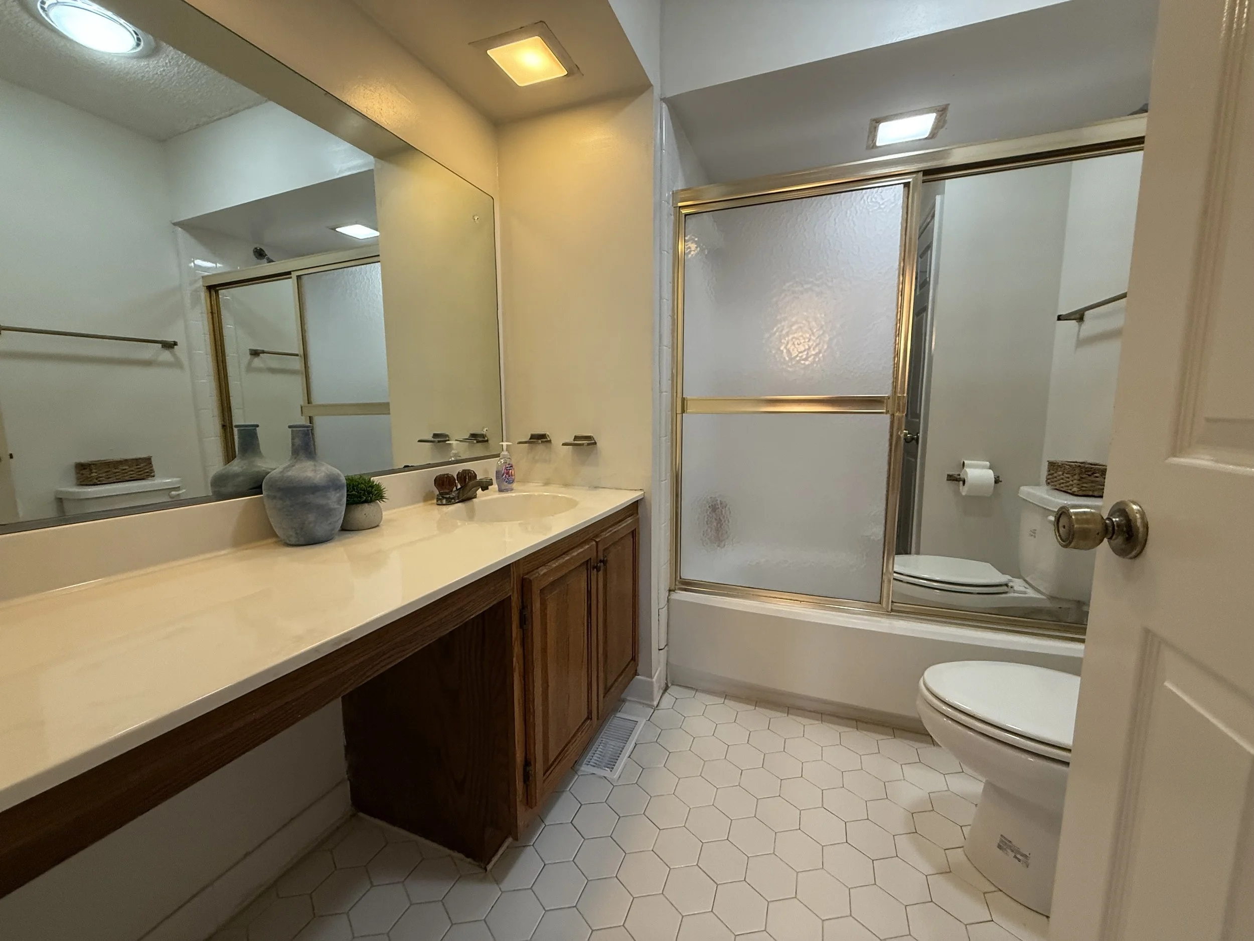 A bathroom with a single sink vanity with wooden cabinets, a large mirror, and decorative vases. To the right is a frosted glass shower door, a toilet, and a small window with a white frame. The floor has small hexagonal tiles.