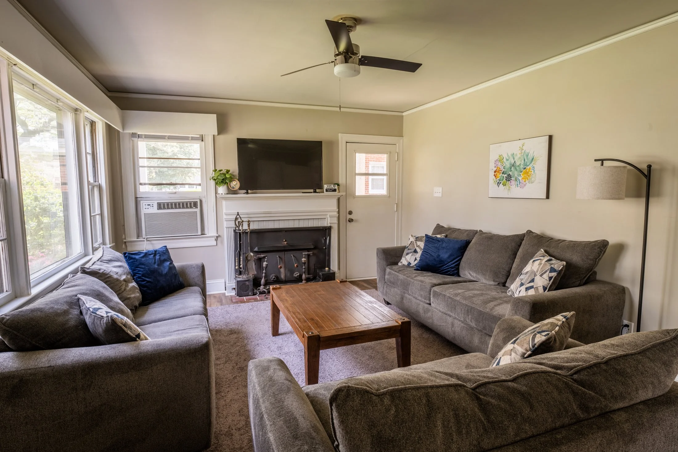 Living room with three gray sofas, adorned with blue and patterned pillows, a wooden coffee table, a fireplace with a television above, windows with a view outside, a wall-mounted lamp, a wall art piece, and a ceiling fan.
