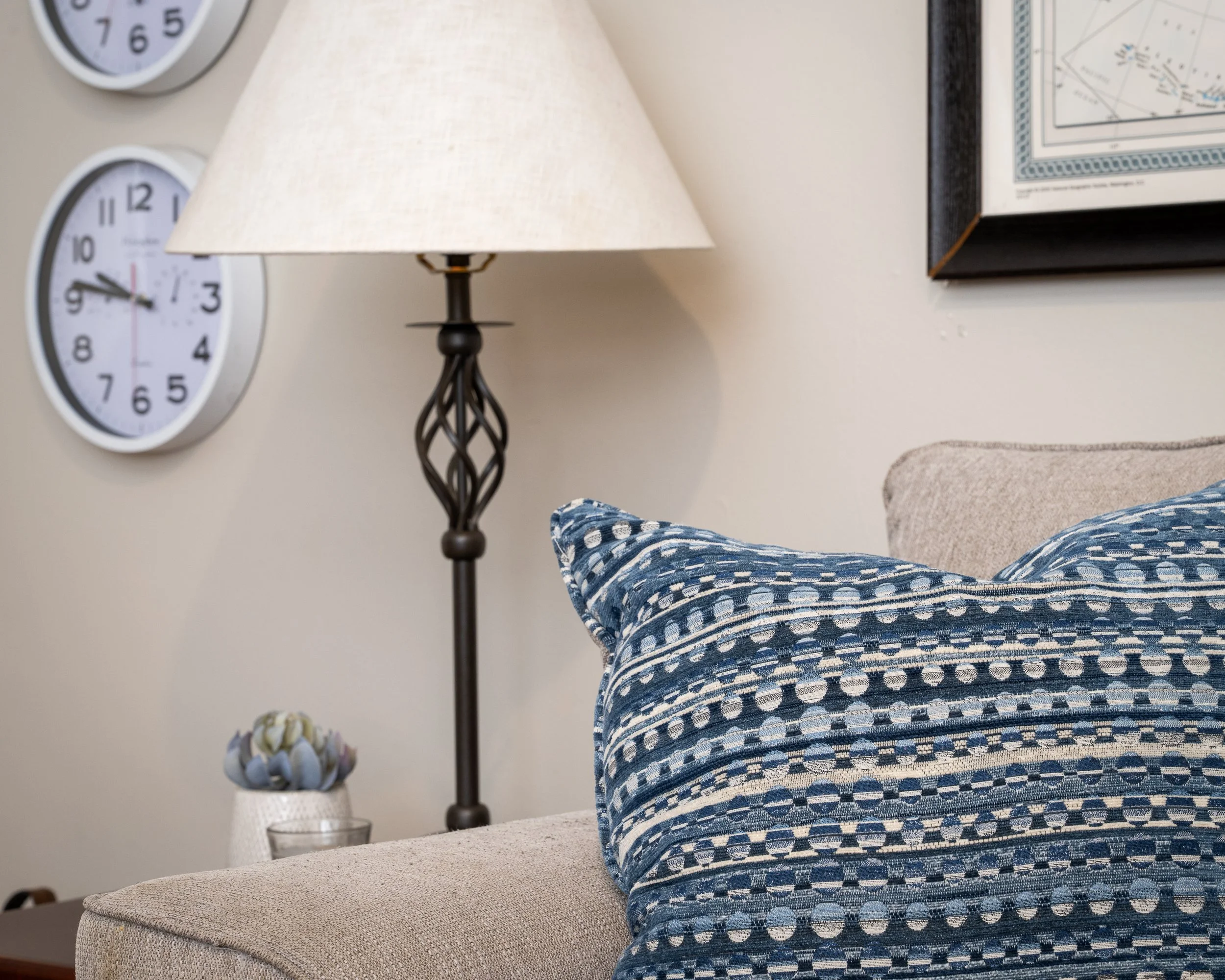 Close-up of a blue and white patterned pillow on a beige couch in a living room, with a side table and decorative items, clocks, a lamp, and framed artwork on the wall.