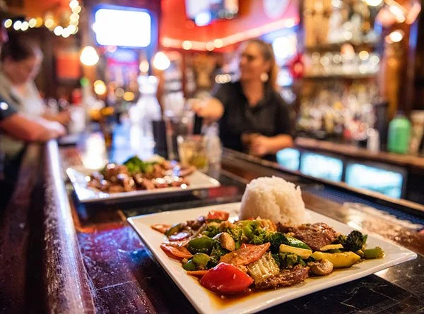 Square white plate with rice and a colorful stir-fry of vegetables and beef on a bar counter in a restaurant. Blurred background with a bartender and a customer.