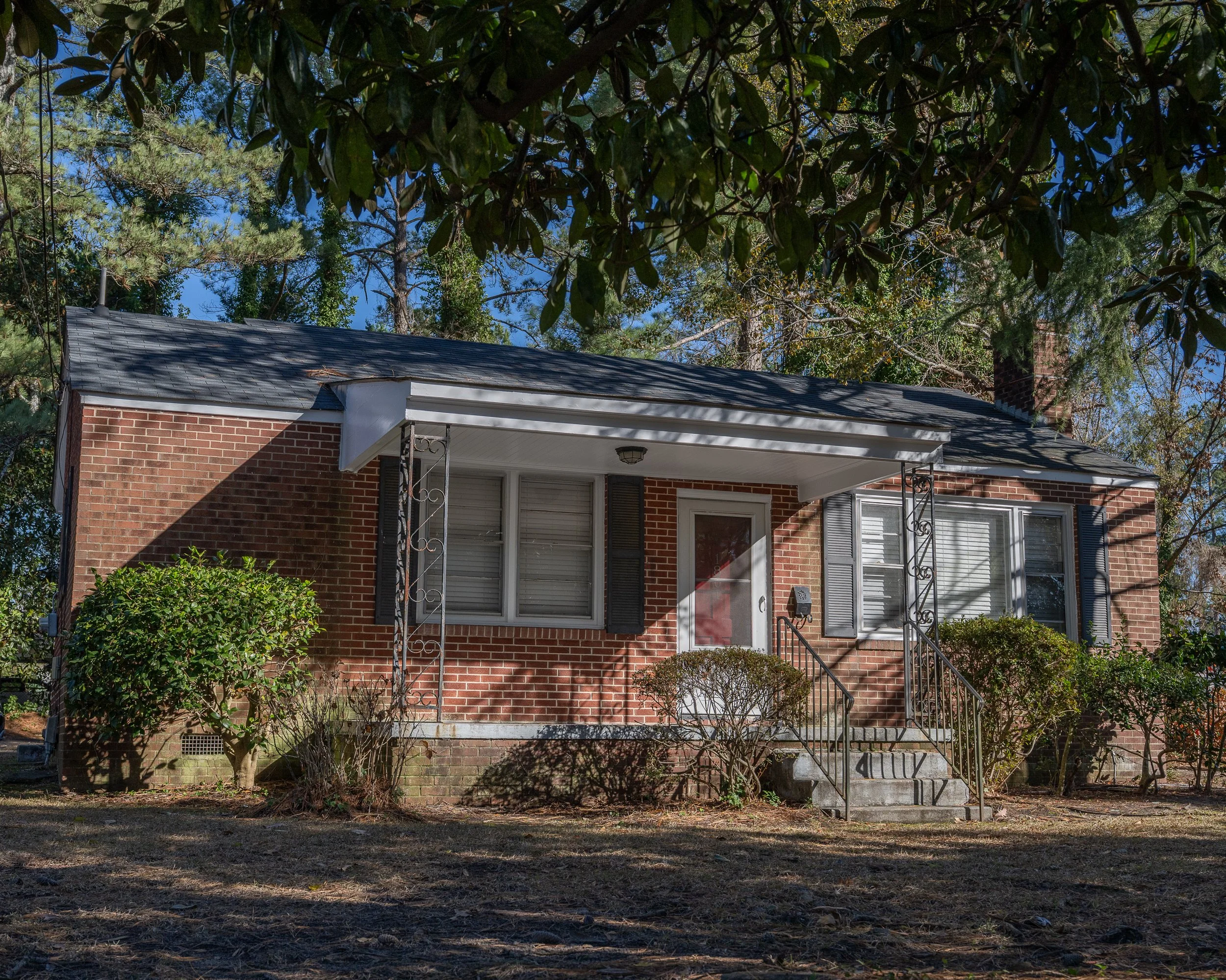 A single-story brick house with a small front porch, steps, and metal railing. Surrounded by bushes and trees, with a lawn in the foreground and clear blue sky in the background.