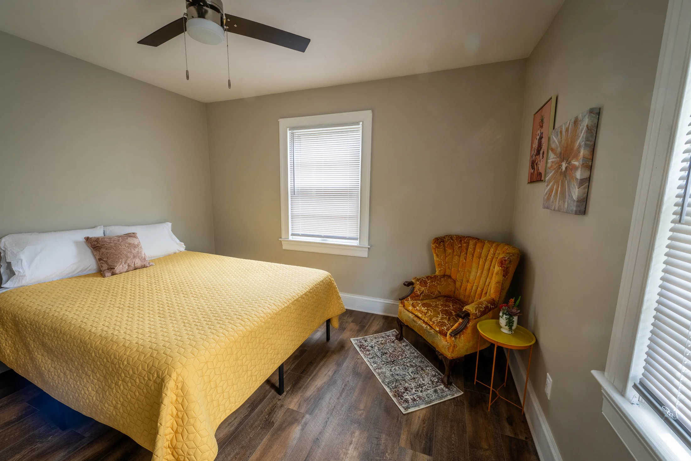 A bedroom with a bed covered in a yellow quilt, a small pink pillow, a vintage orange armchair with wooden accents, a yellow side table with a potted plant, a window with blinds, hardwood floors, and three framed wall art pieces.