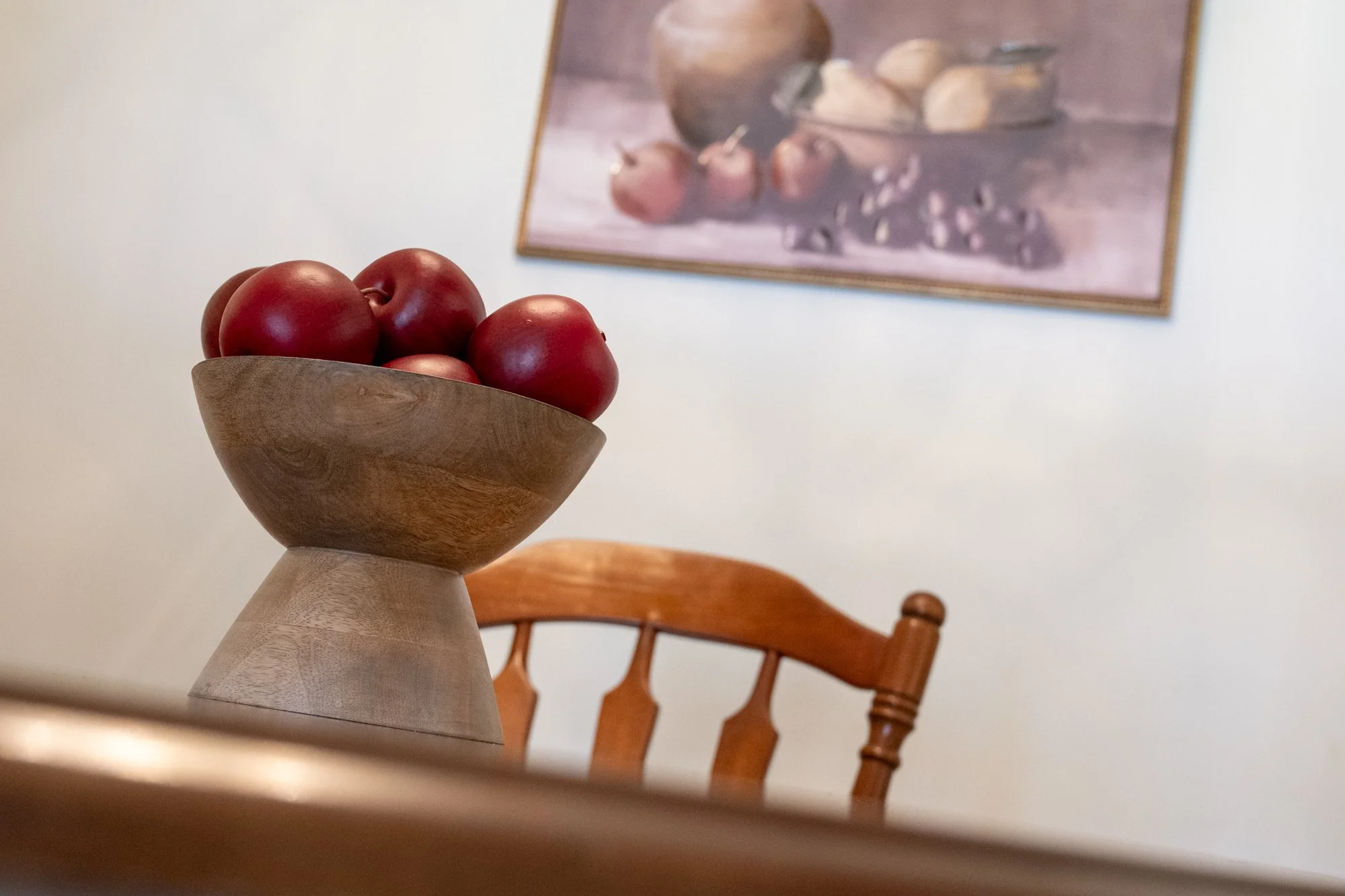 A wooden bowl filled with red apples on a wooden table in a room with a wooden chair and a framed painting of fruits on the wall.