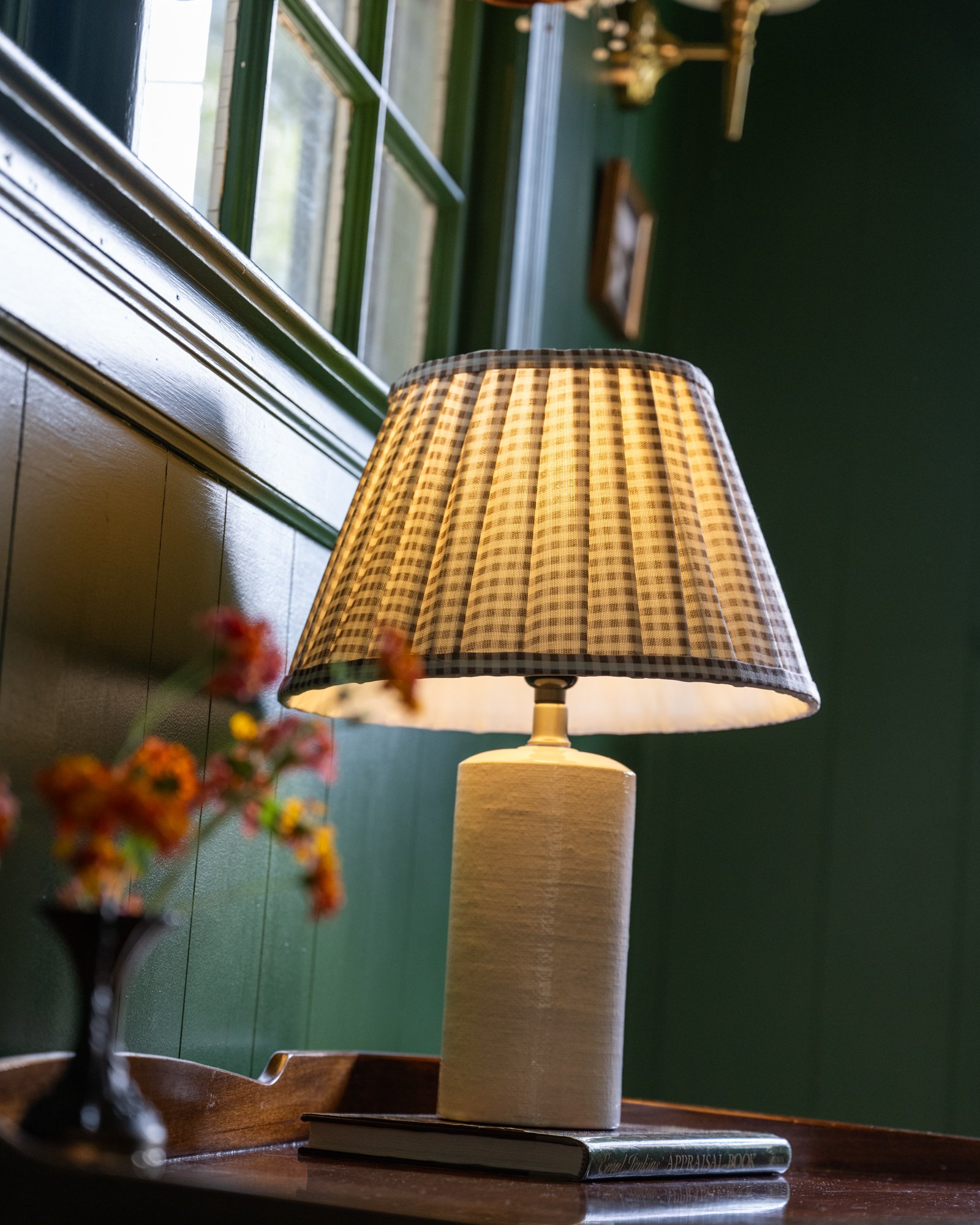 Table lamp with checkered lampshade on a wooden table next to a small vase of flowers, in a windowed room with green walls.