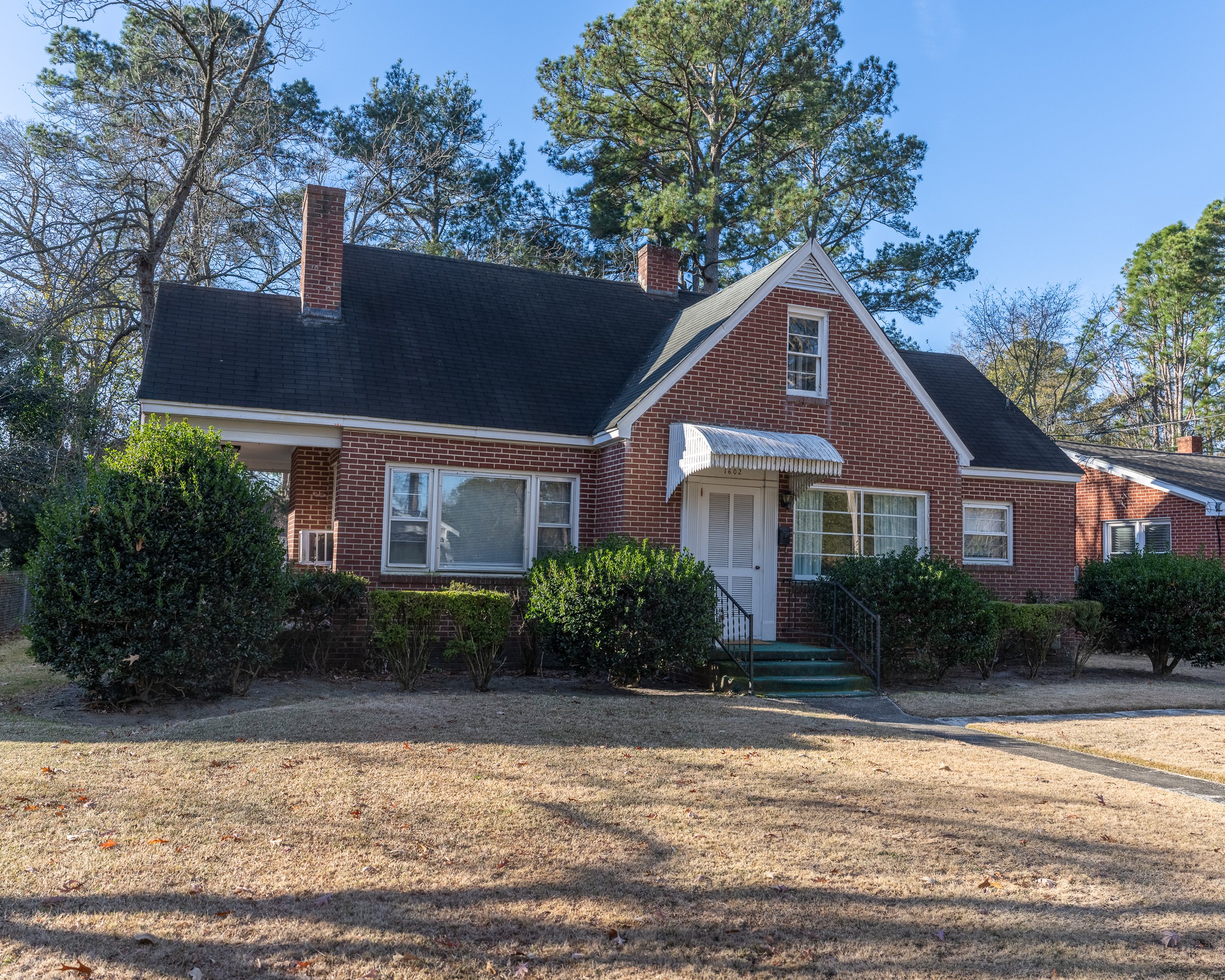 A brick house with a black roof and front porch, surrounded by bushes and a lawn, under a clear blue sky.