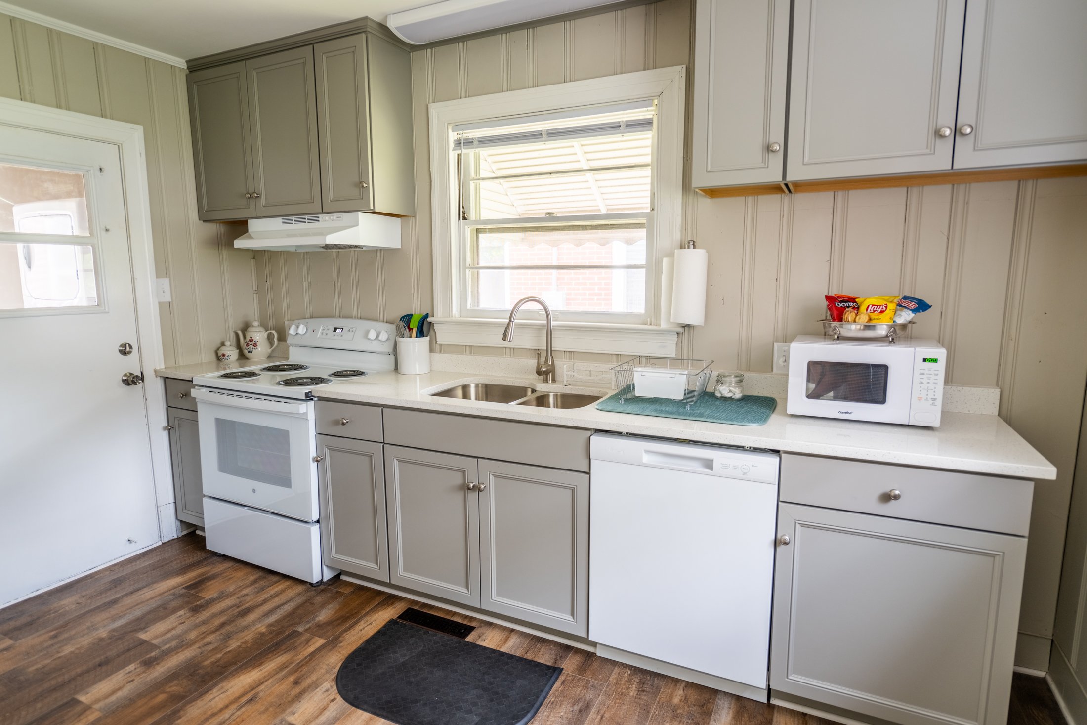 Kitchen with beige cabinets, white appliances including a stove, microwave, and dishwasher, a window above the sink, and a wooden floor.
