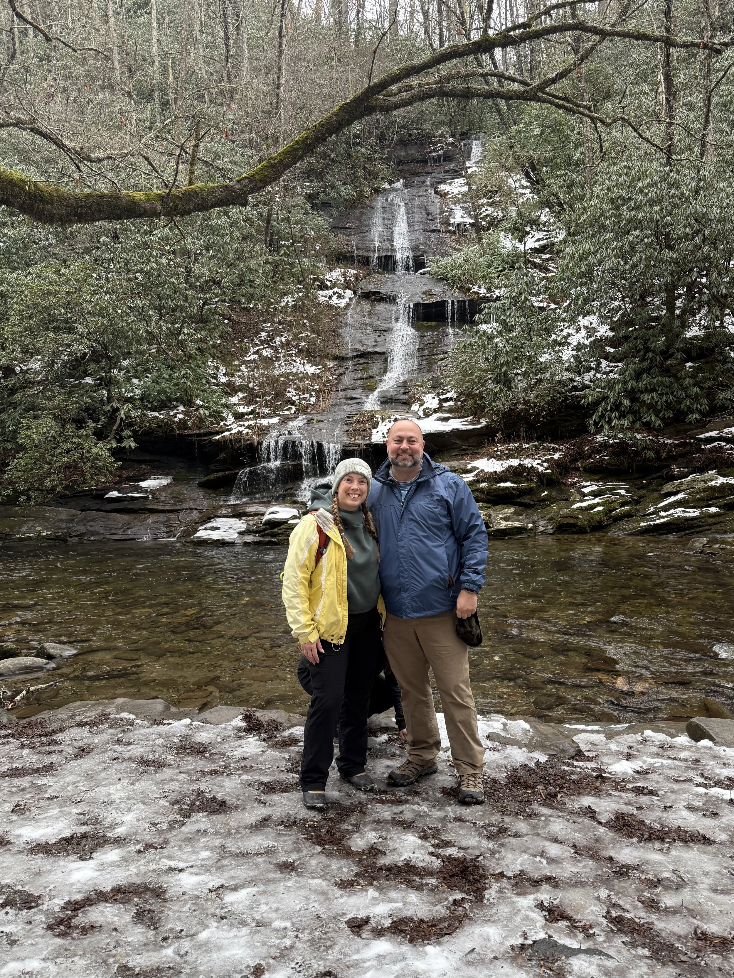 A couple standing on a snow-covered ground by a river with a waterfall in the background in a wooded area.