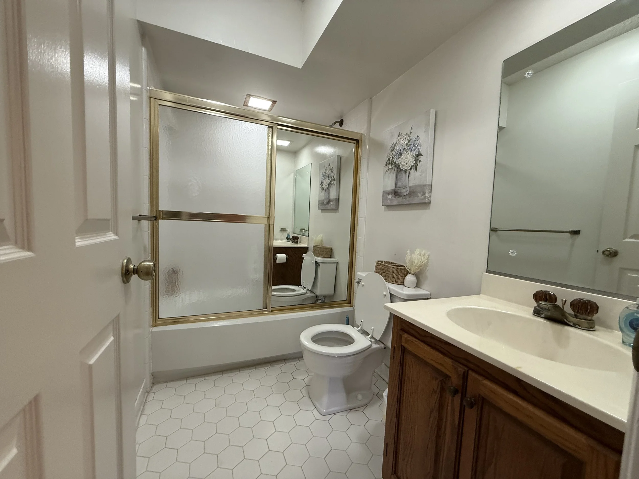A small bathroom with a vanity, sink, shower with a frosted glass door, toilet, and framed pictures of flowers on the wall.