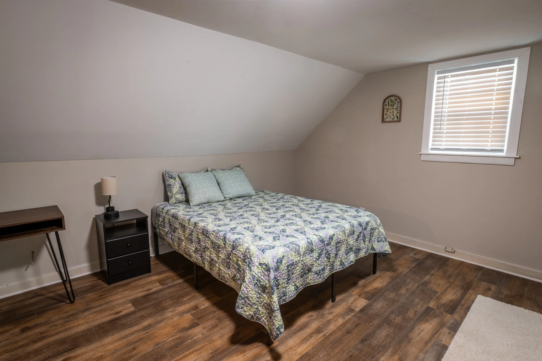 A bedroom with a bed, a black nightstand with two drawers, a small lamp, a wooden desk with black hairpin legs, and a window with white blinds. The room has hardwood flooring and a sloped ceiling.