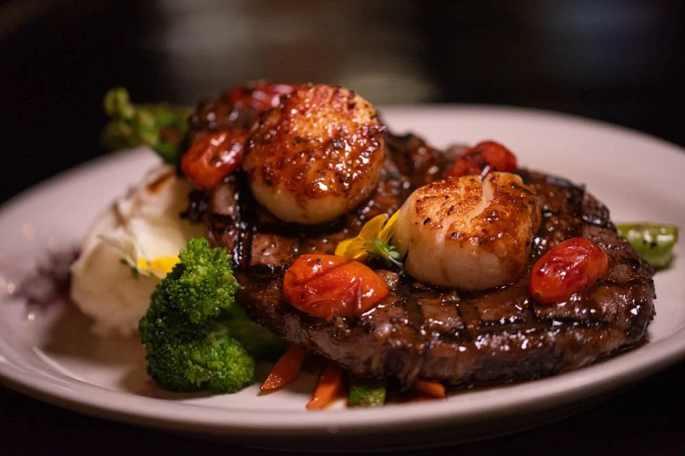 A plate with grilled steak topped with scallops, cherry tomatoes, and garnished with vegetables like broccoli, carrots, and cauliflower.