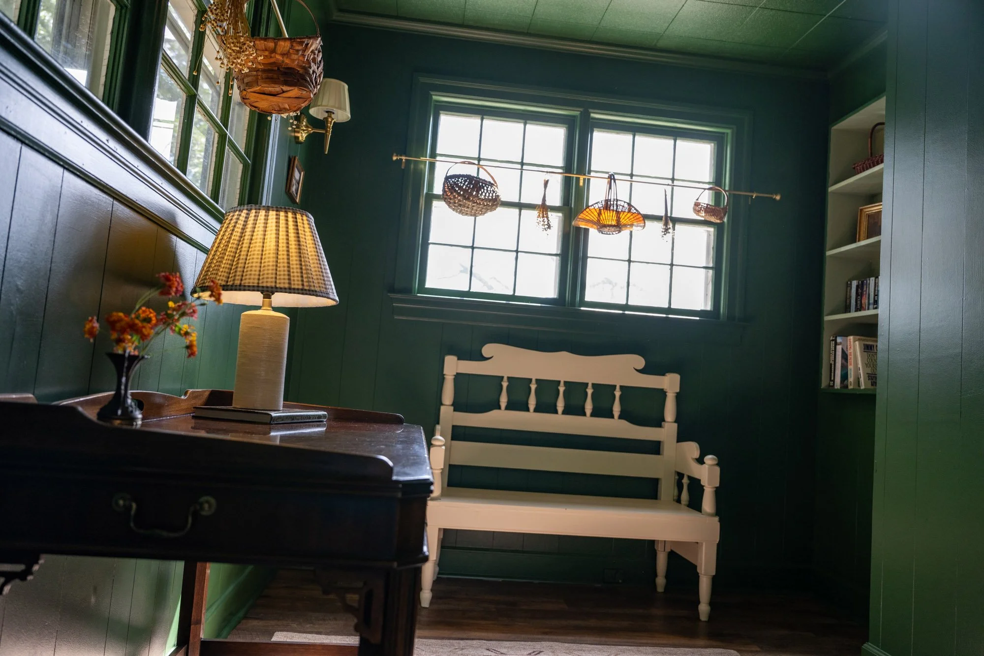 Interior of a cozy room with green walls, a window with multiple panes, and a white bench. A table with a lamp, a vase of flowers, and books is in the foreground. There are hanging baskets and woven decor near the window, and a bookshelf on the right side.