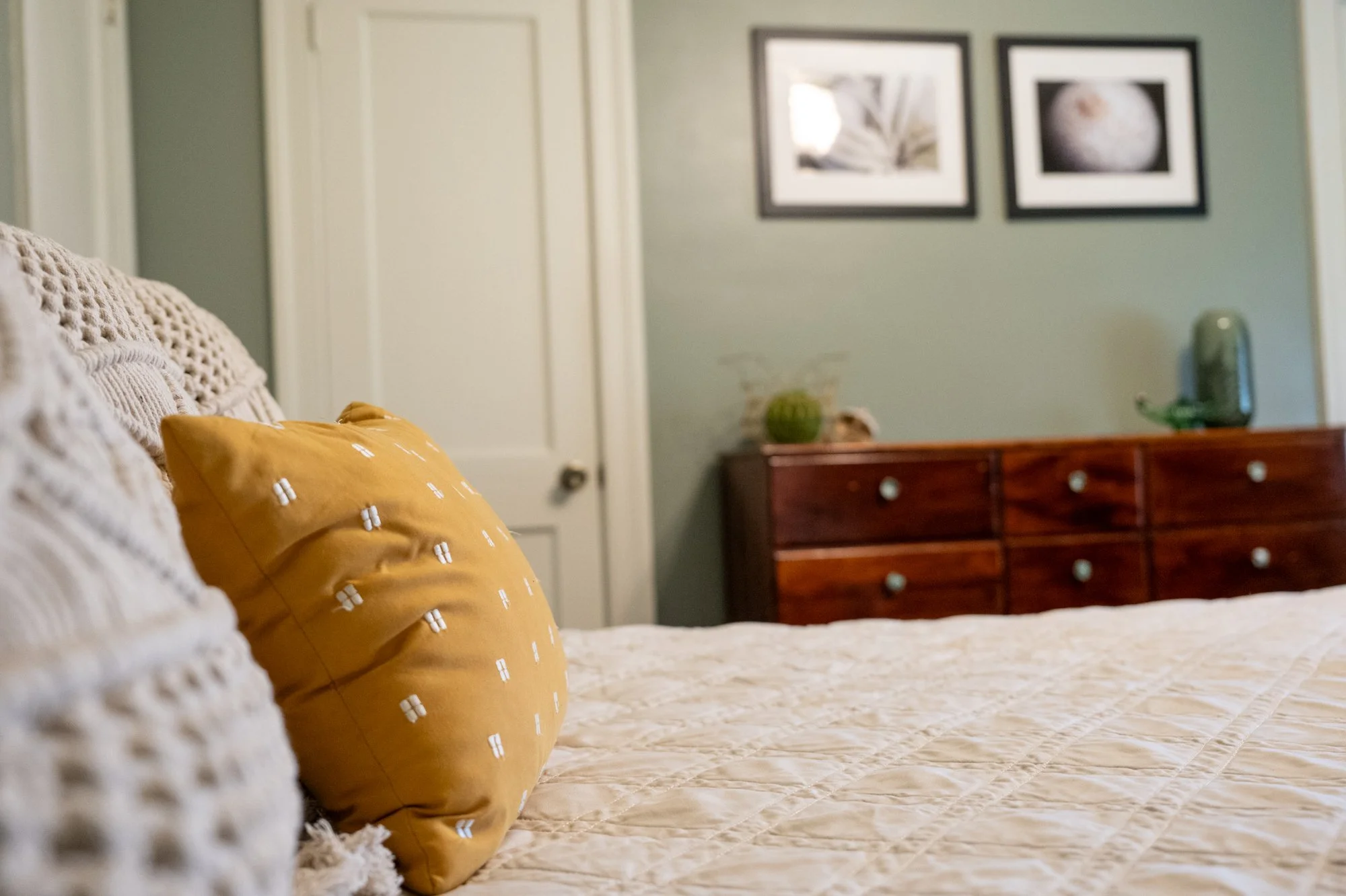 A cozy bedroom with a bed covered in a beige quilt and a decorative yellow pillow with white embroidered details. In the background, a wooden dresser and framed black-and-white photographs of nature on the wall are visible.