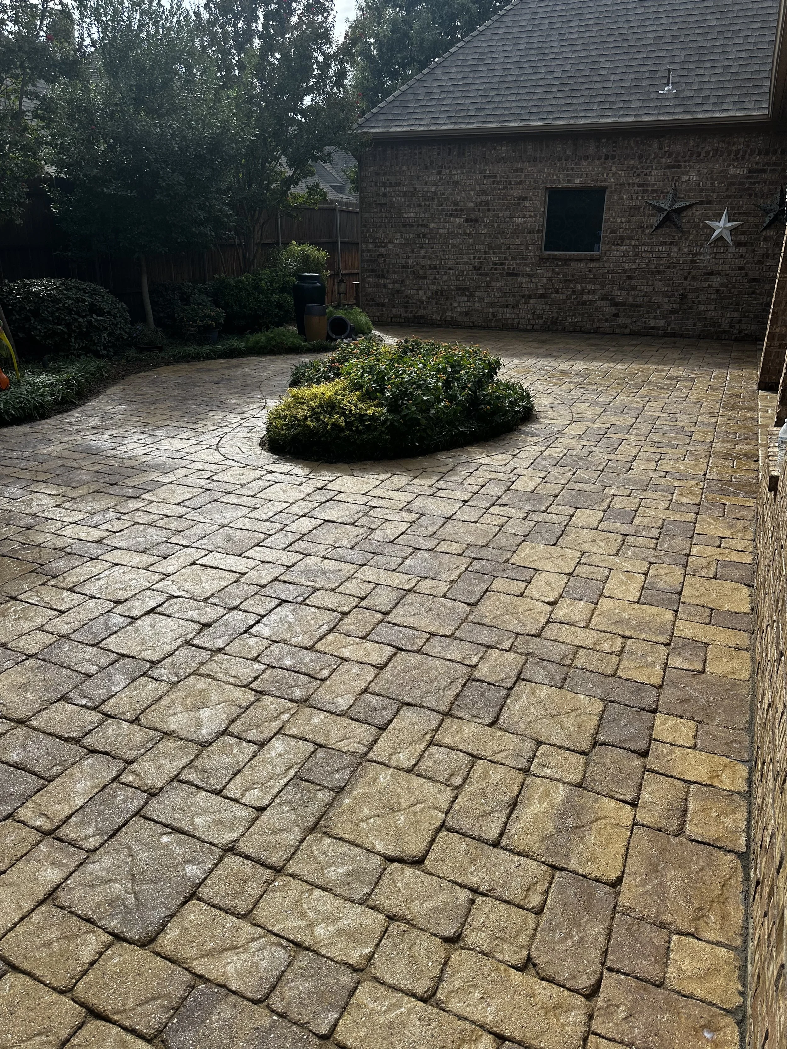 Clean, paved backyard with stone bricks, brick house wall on the right, green bushes, trees, and decorative stars on the house wall.