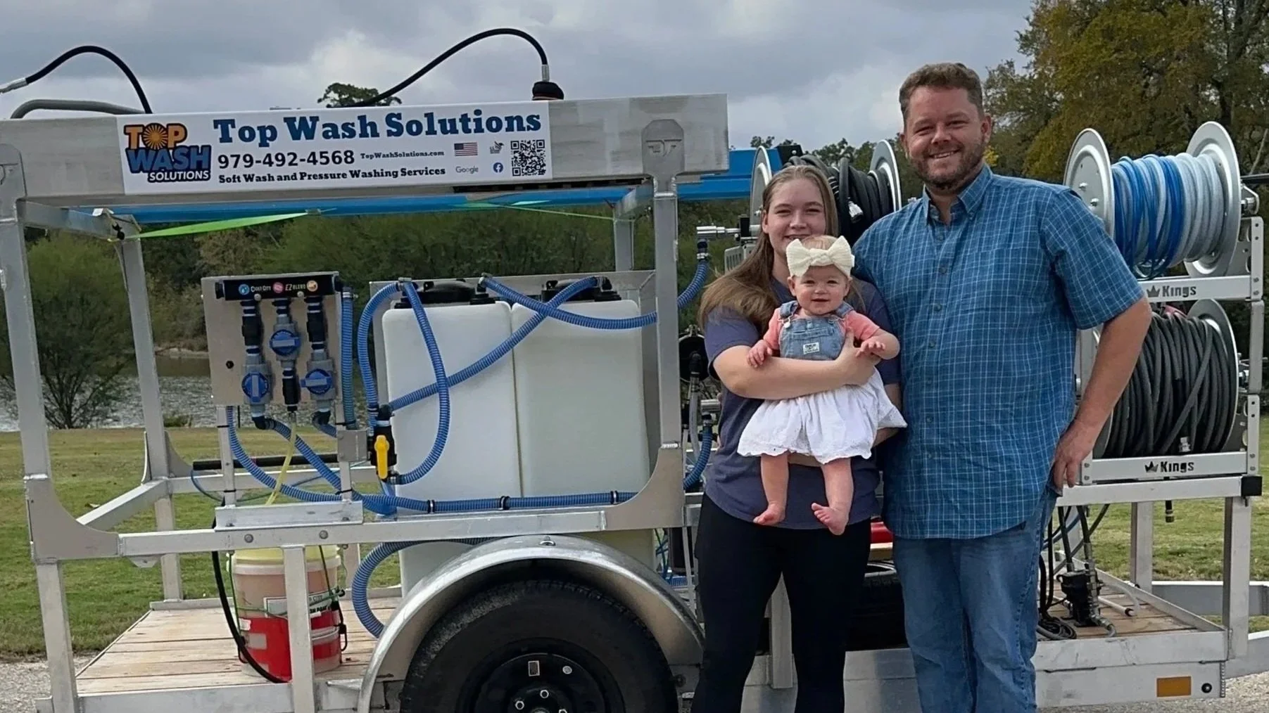 A family standing in front of a mobile pressure washing trailer, with a woman, a man, and a baby girl smiling outdoors near a river, trees, and cloudy sky.