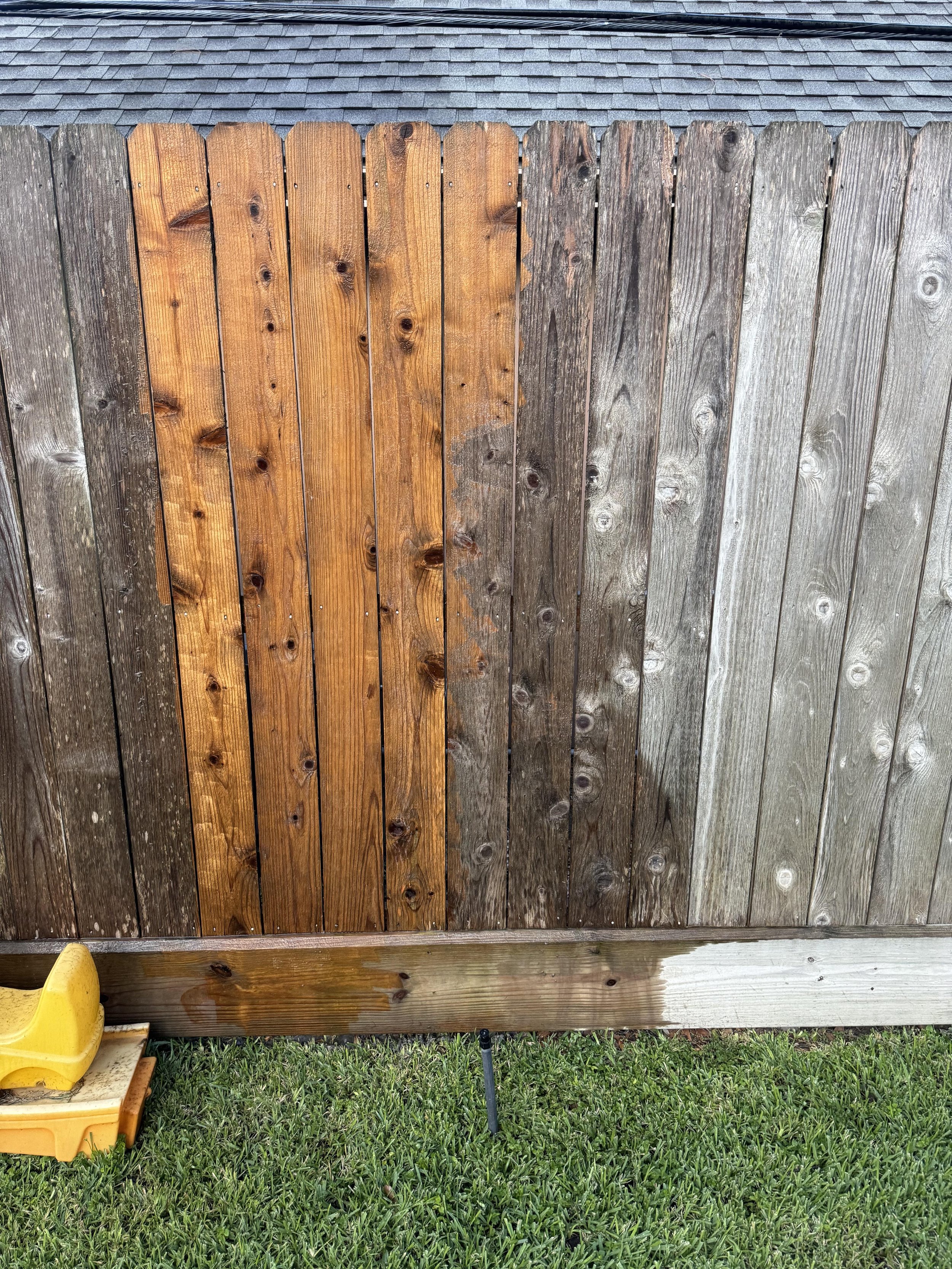 A wooden fence with alternating weathered and stained panels, set on a grassy lawn with a yellow children's sandbox in the lower left corner.