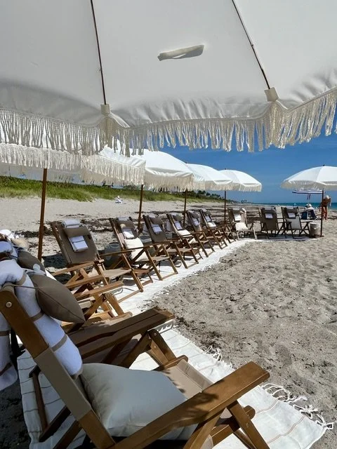 Beach chairs with white towels under large white beach umbrellas on a sandy shore.