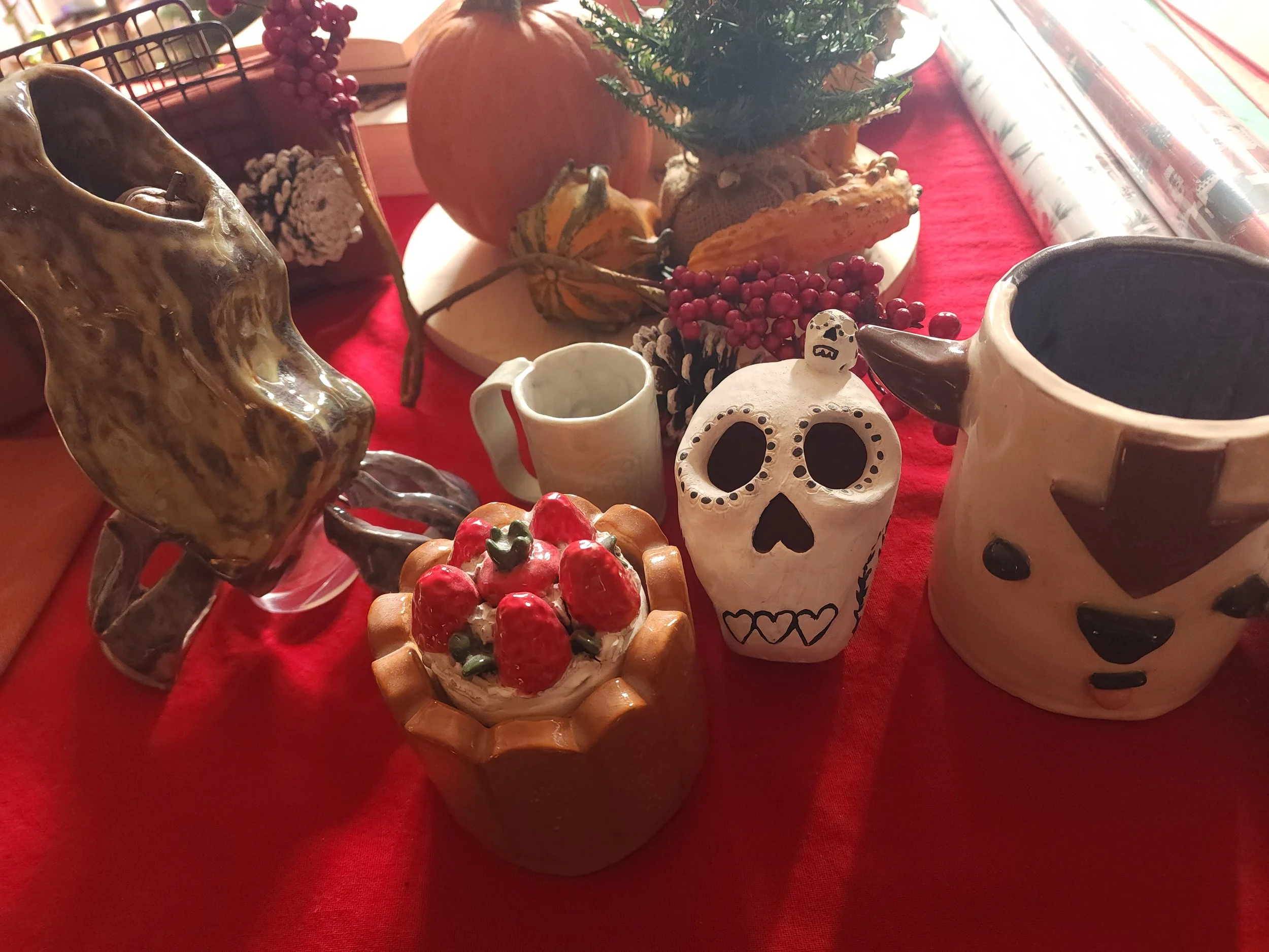 Decorative table with ceramic skull, pumpkin, small Christmas tree, mug, and other festive items on a red tablecloth.