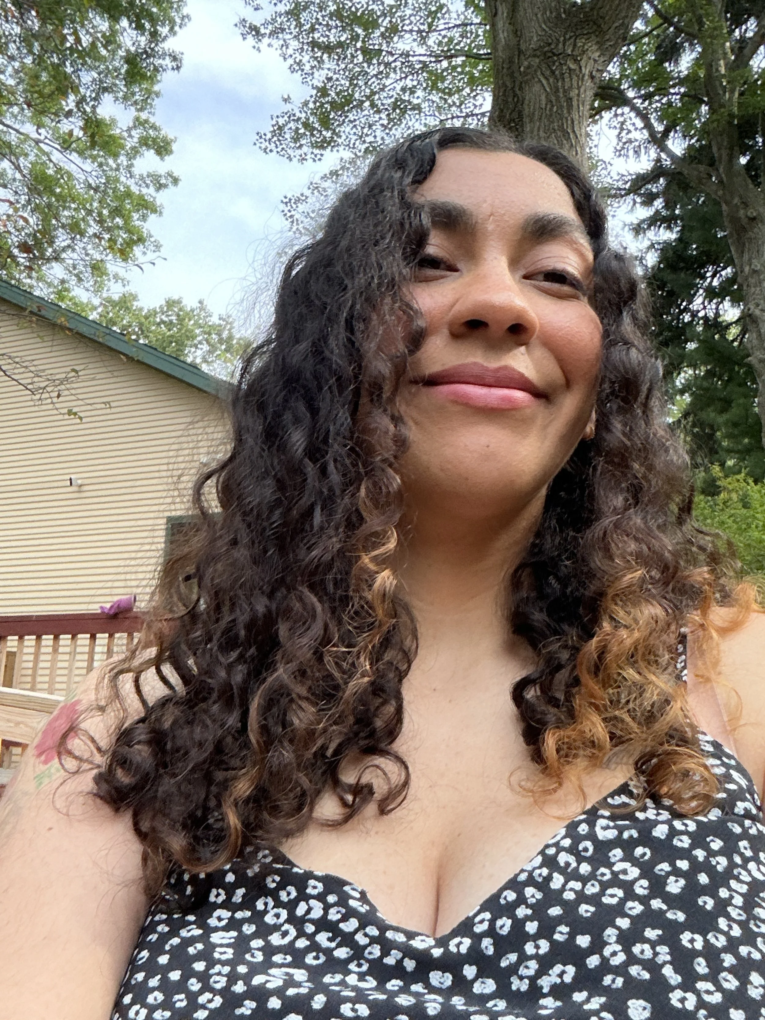 A woman with long curly dark hair smiling outdoors, wearing a black and white patterned top, with trees and a house in the background.