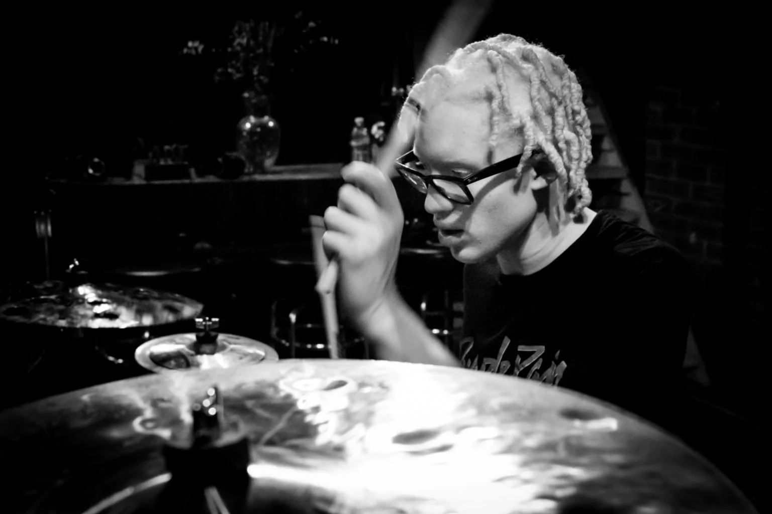 A person with light-colored dreadlocks and glasses playing a drum set in a dimly lit room.