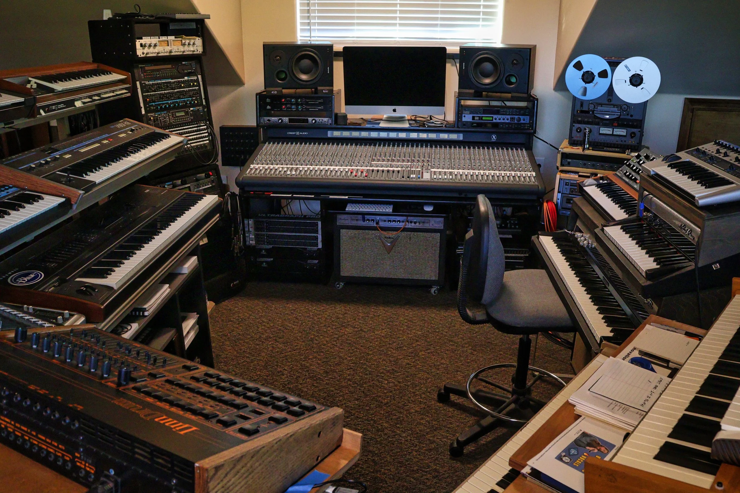 A music studio with various electronic musical instruments and equipment, including keyboards, synthesizers, mixing console, studio monitor speakers, tape reels, and audio gear, arranged in a room with a window.