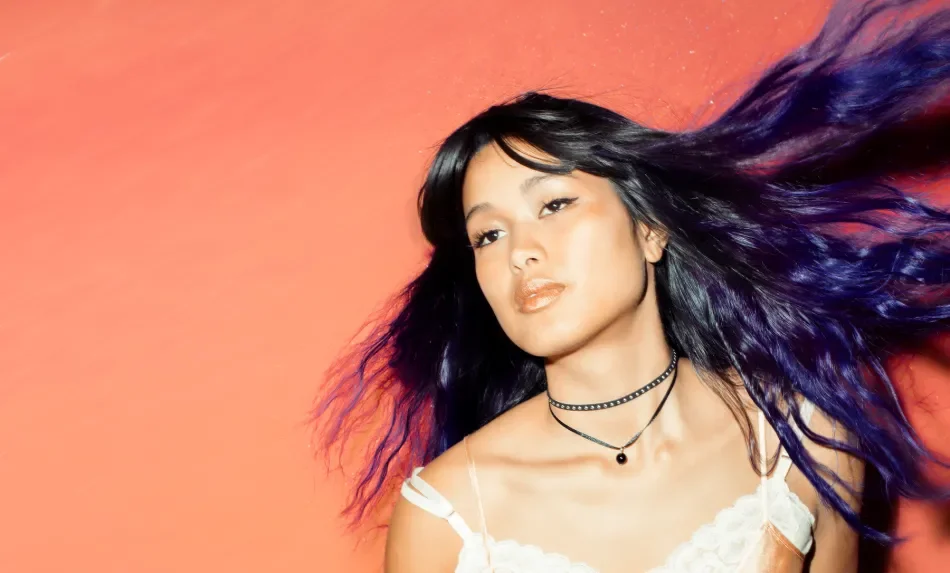 Young woman with long, wavy black and purple hair posing against an orange background, wearing a white spaghetti strap top and black layered choker necklaces.