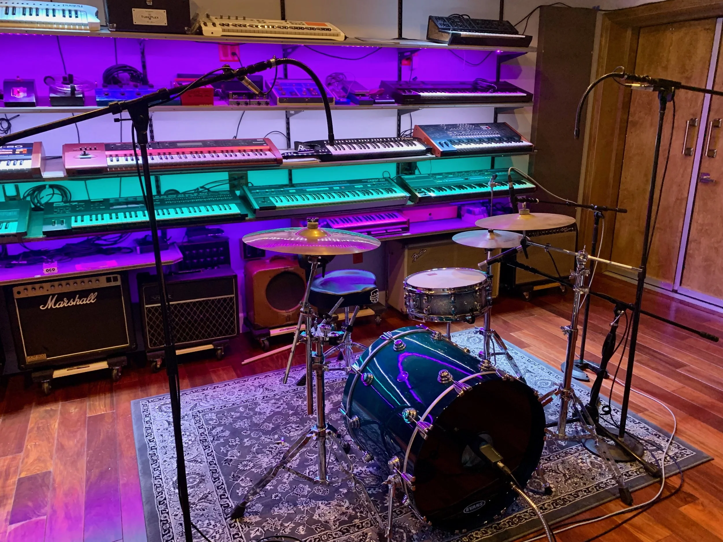 Music studio with various keyboards, microphones, drums, and amplifiers, illuminated by purple and blue lighting, on a wooden floor with a patterned rug.