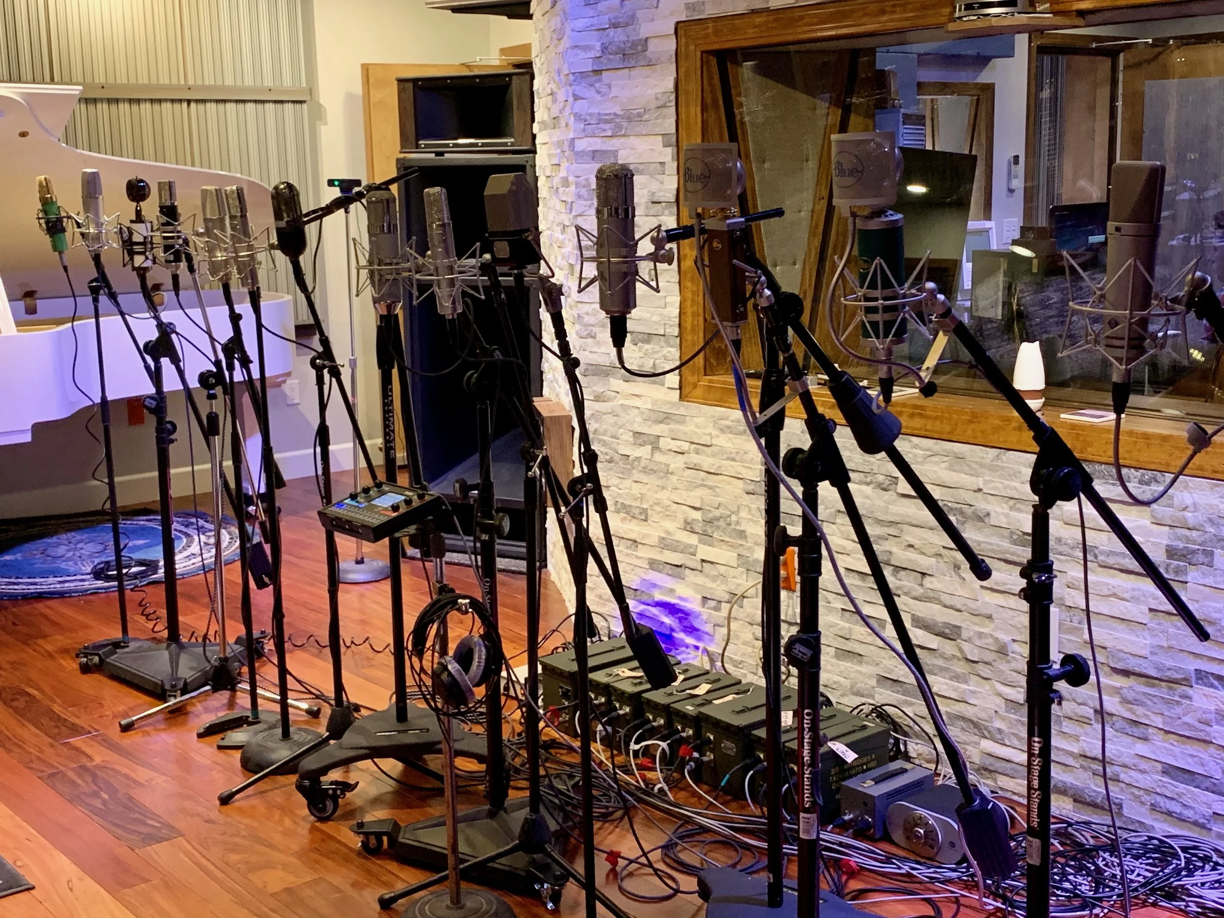 A recording studio with multiple microphones on stands set up in front of a mixing console. The room has a wooden floor, a stone accent wall, and a large window looking into an adjacent control room where audio equipment is visible.