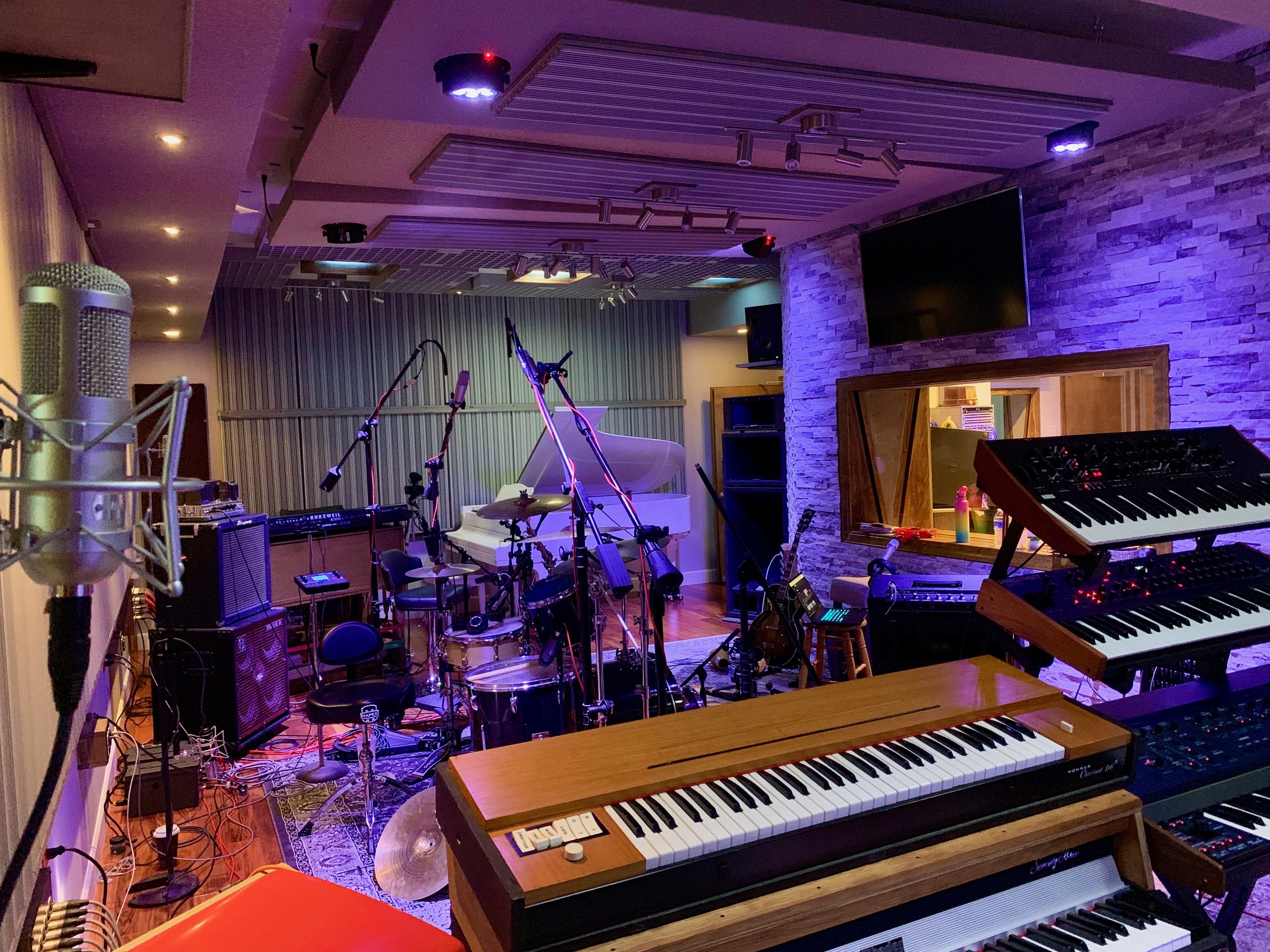 Music studio with various instruments, including a grand piano, electronic keyboard, drums, and microphones, illuminated with purple and warm lighting.