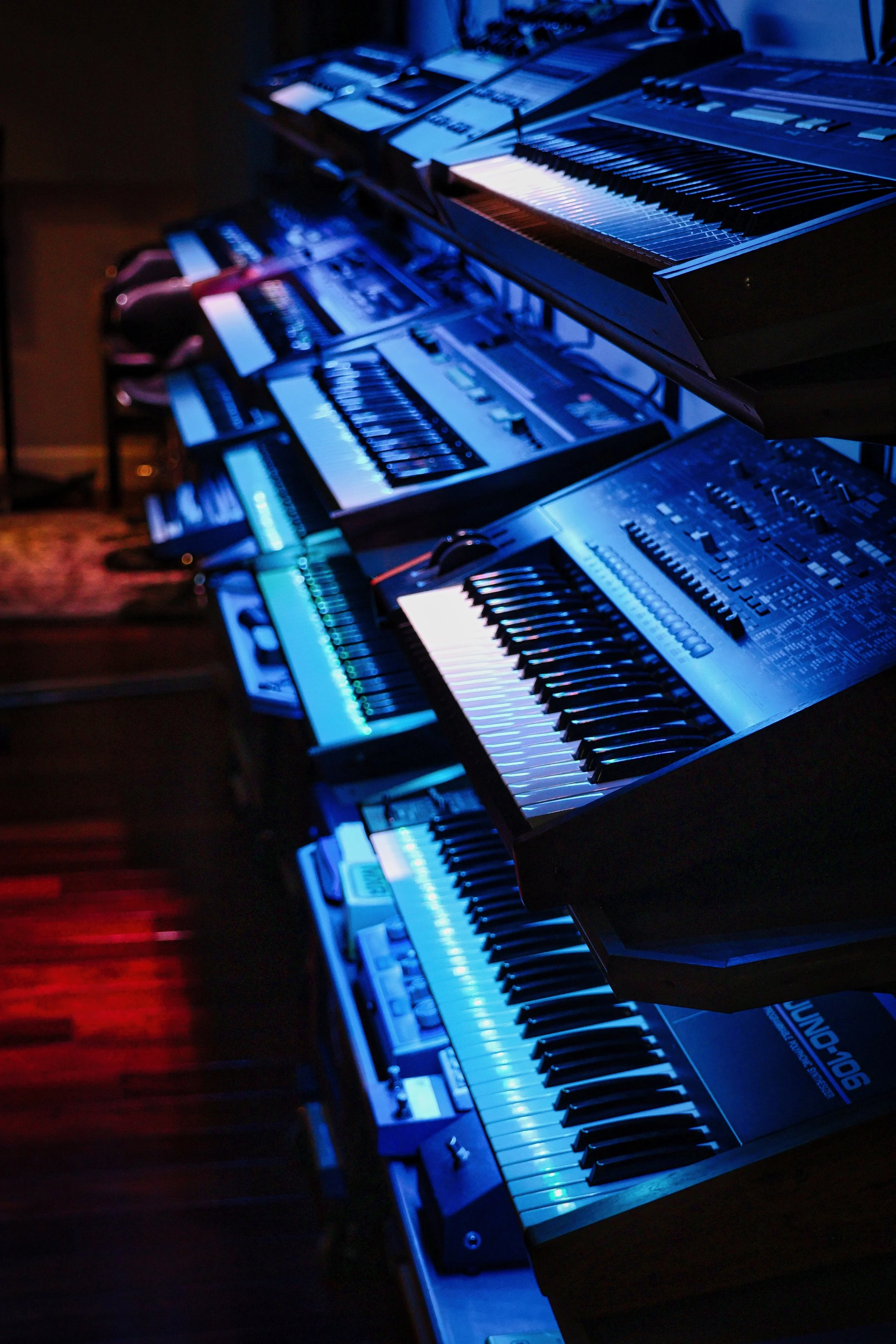 Multiple electronic keyboards stacked on a stand, illuminated with blue and red stage lighting, in a dark setting.