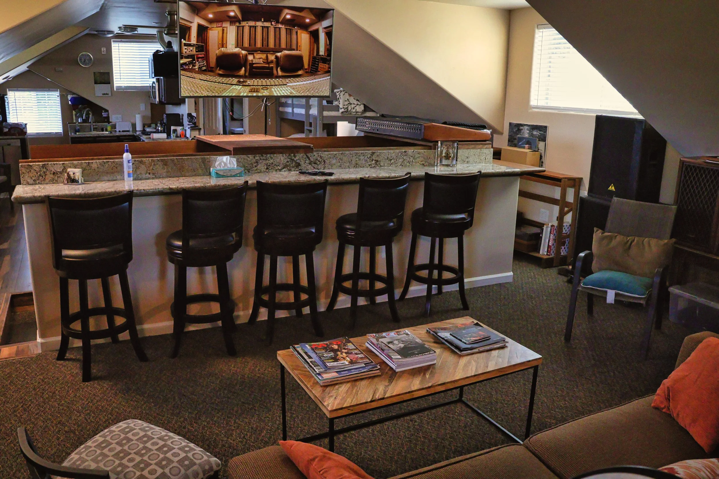 A cozy attic music studio with a large monitor displaying a recording studio interface, a granite countertop with four black bar stools, a small wooden table with magazines, a drum switcher on the countertop, and a window with blinds allowing natural