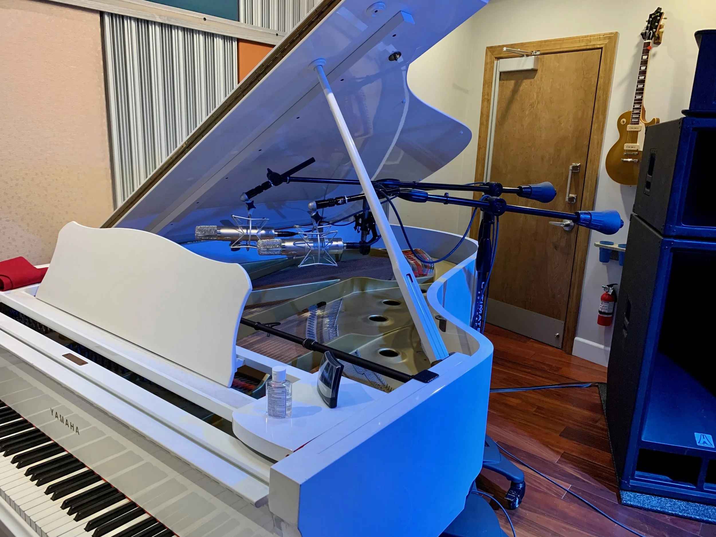 White Yamaha grand piano with its lid open, music stand inside, black and white keys visible, placed in a room with wooden floor, brown door, guitar hanging on the wall, and a blue speaker cabinet.