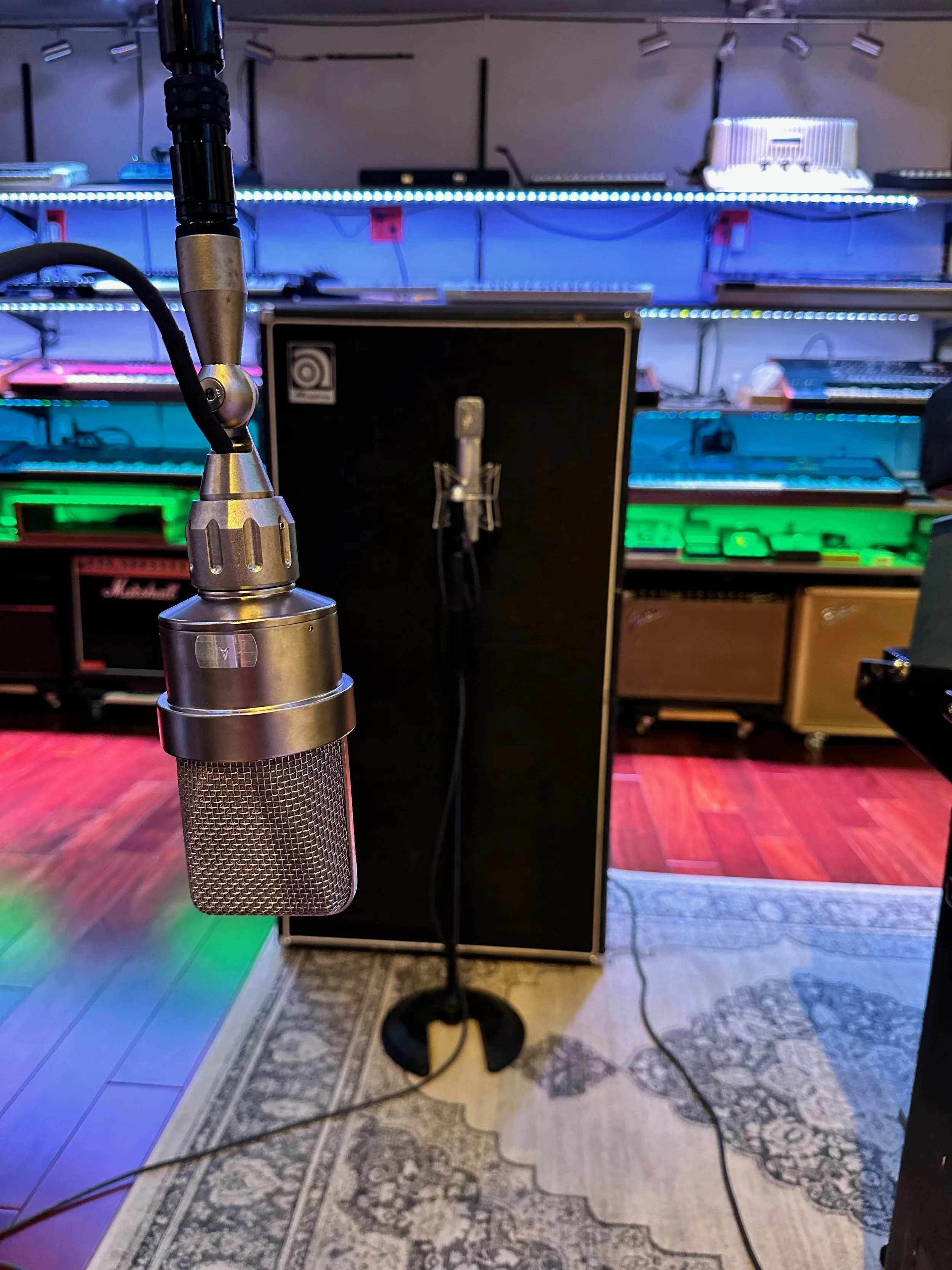 A professional recording studio featuring a vintage-style microphone on a stand, with audio equipment and colorful lighting in the background.
