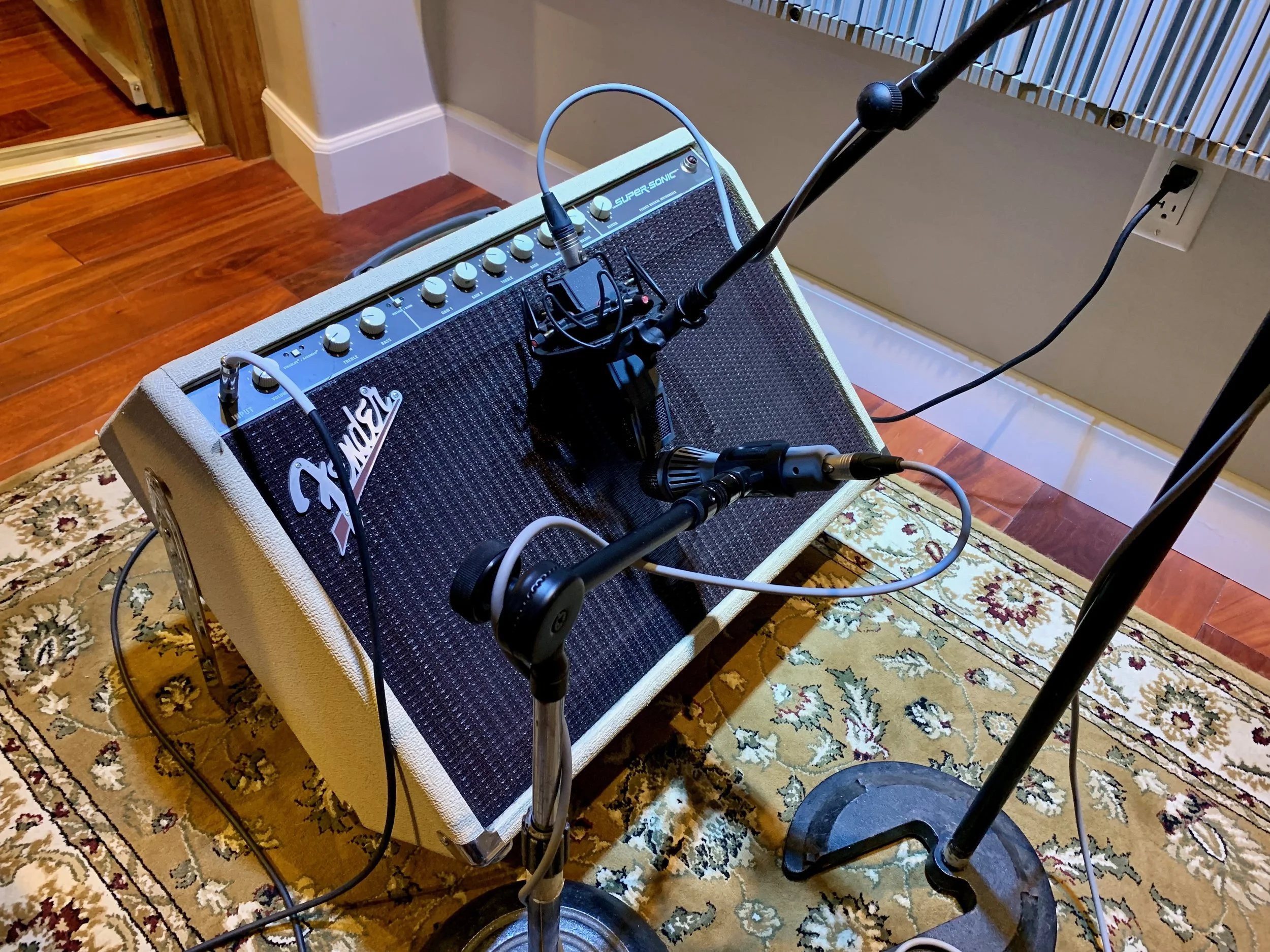 Guitar amplifier on a patterned rug with a microphone stand and microphone in front, connected to the amplifier, near a wall with an electrical outlet.