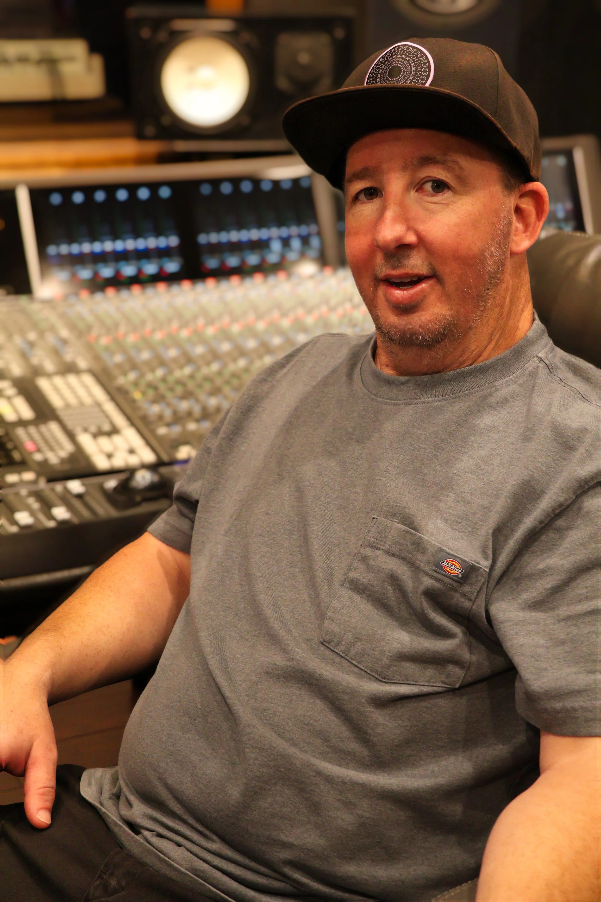 A man wearing a gray Dickies t-shirt and a black baseball cap, sitting in a recording studio with sound equipment and mixing console in the background.
