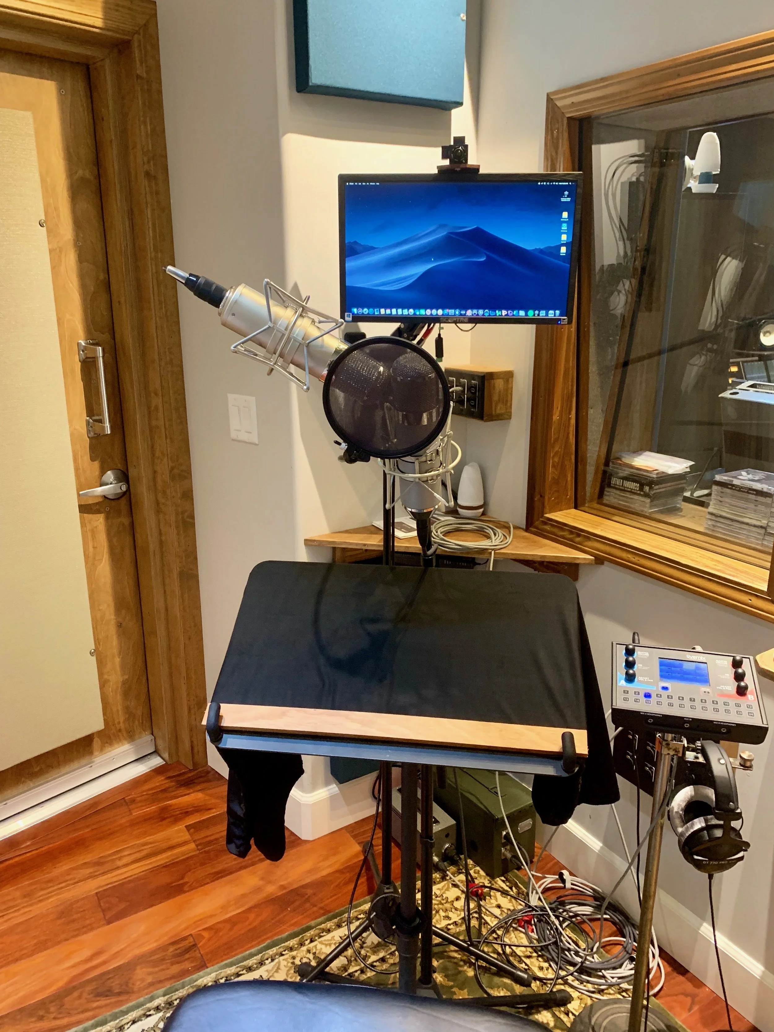 Recording studio setup with a microphone, pop filter, computer monitor, audio equipment, and headphones in a room with hardwood floors.