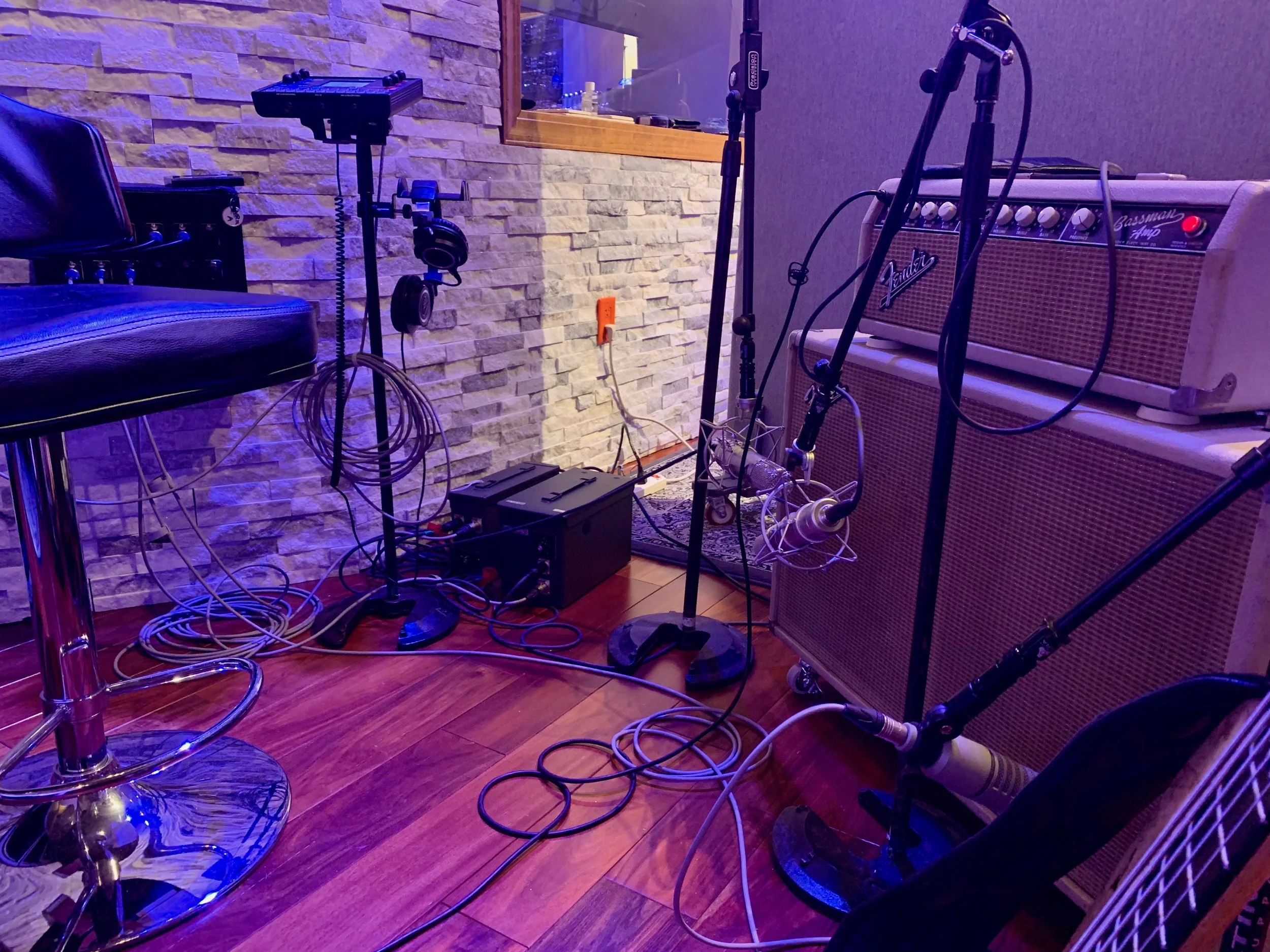 Recording studio with musical equipment including a bass amplifier, microphone stands, headphones, and a chair, set against a stone wall and wooden floor.