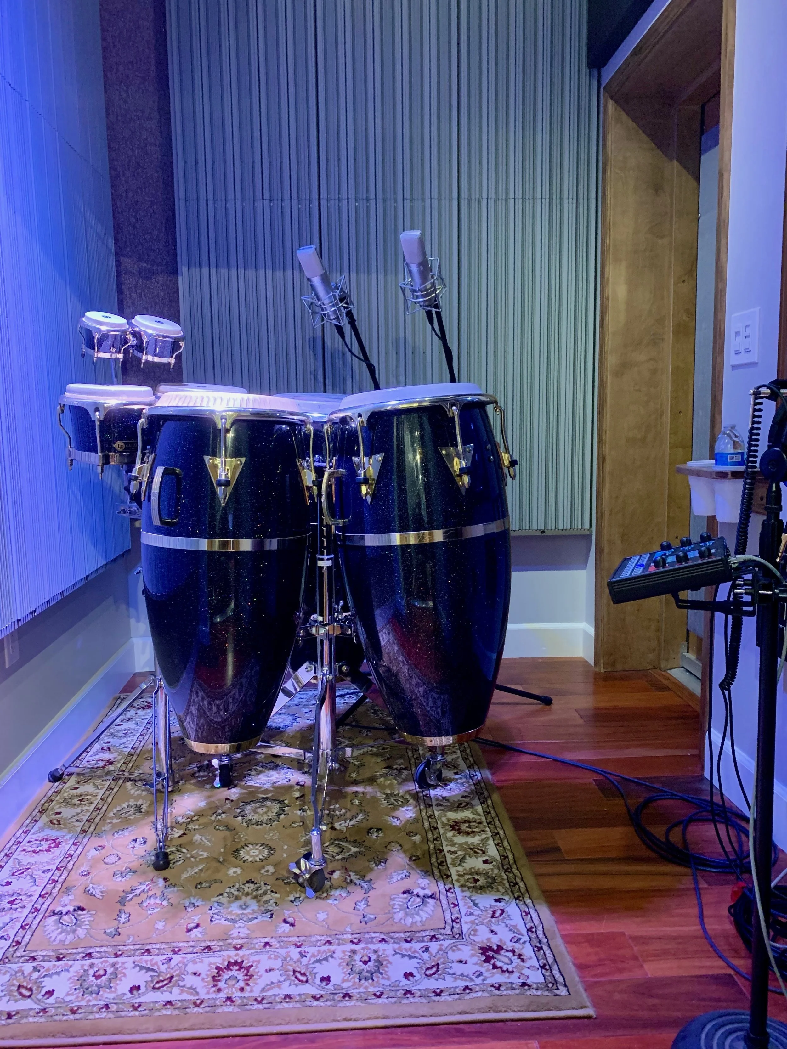 A music recording studio features a set of conga drums on a colorful bashkir rug, two studio microphones on stands, and various audio equipment on the right side in a well-lit room with wood and acoustic panel walls.
