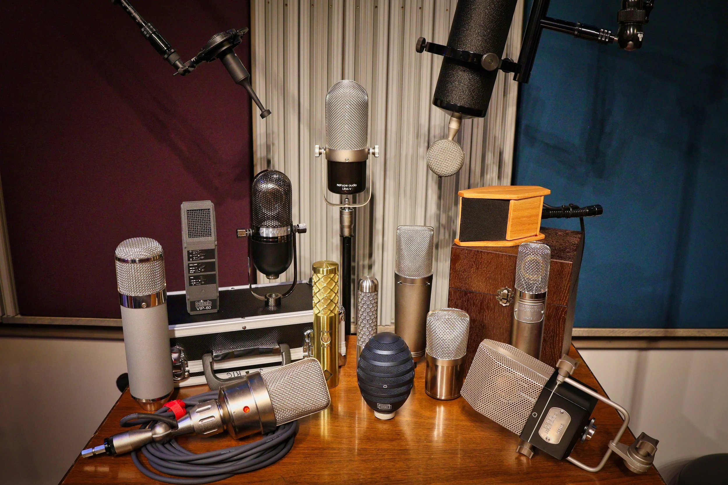 Multiple microphones of various types and sizes arranged on a wooden table in a recording or studio setting.