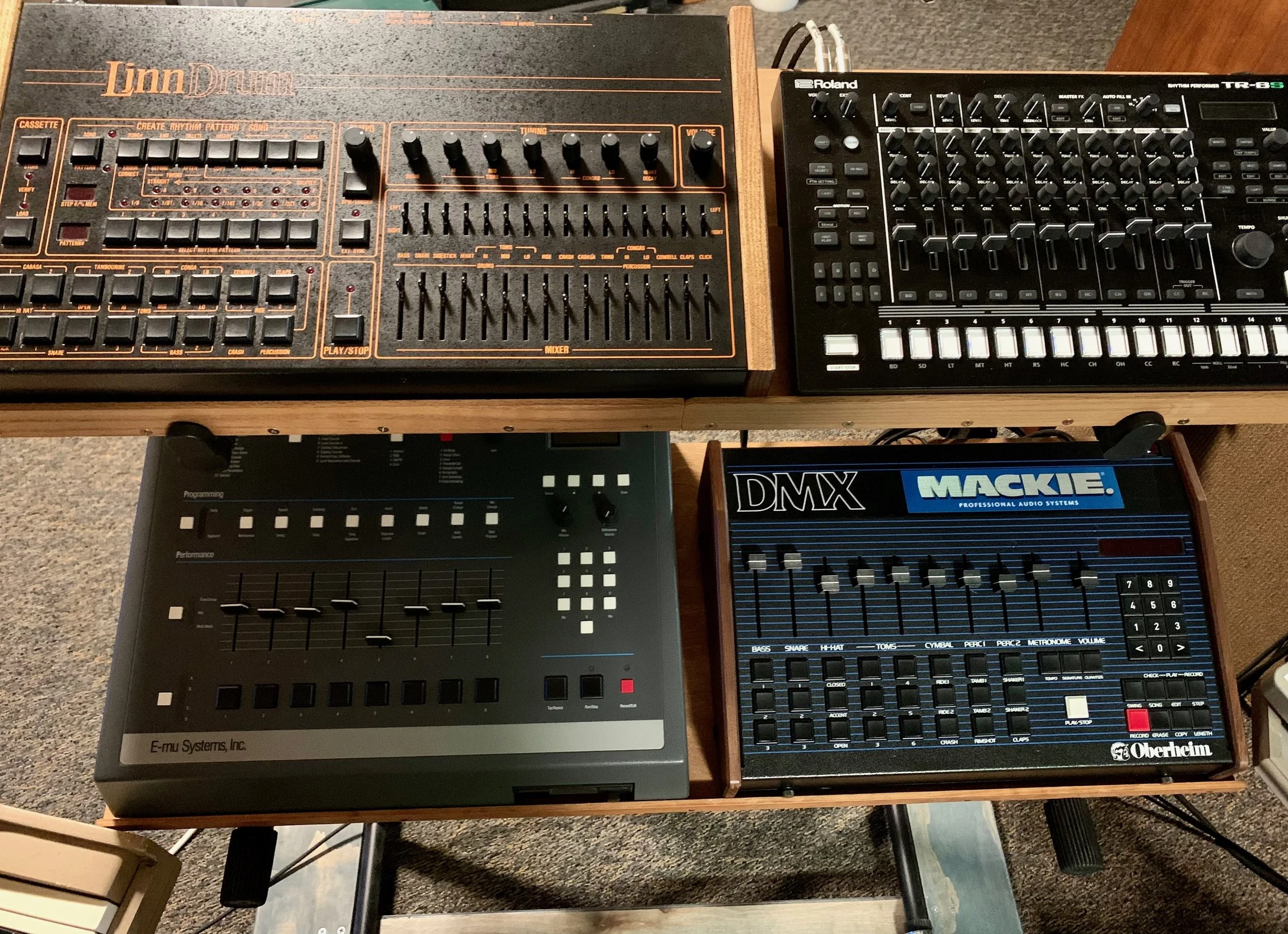 Photo of three electronic music synthesizers and drum machines placed on a wooden table. The devices include a LinnDrum drum machine, a Roland TR-8S drum machine, and an Oberheim drum machine. Various knobs, buttons, faders, and digital displays are 
