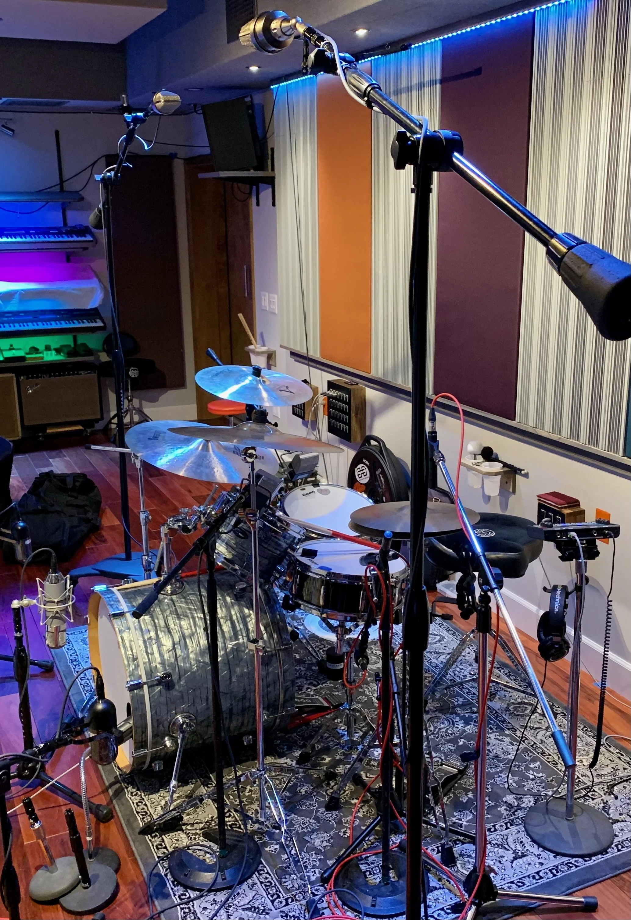 A recording studio room with musical instruments, including drums, a keyboard, microphone stands, and various audio equipment. The room has colorful lighting with blue accents and patterned wall panels.