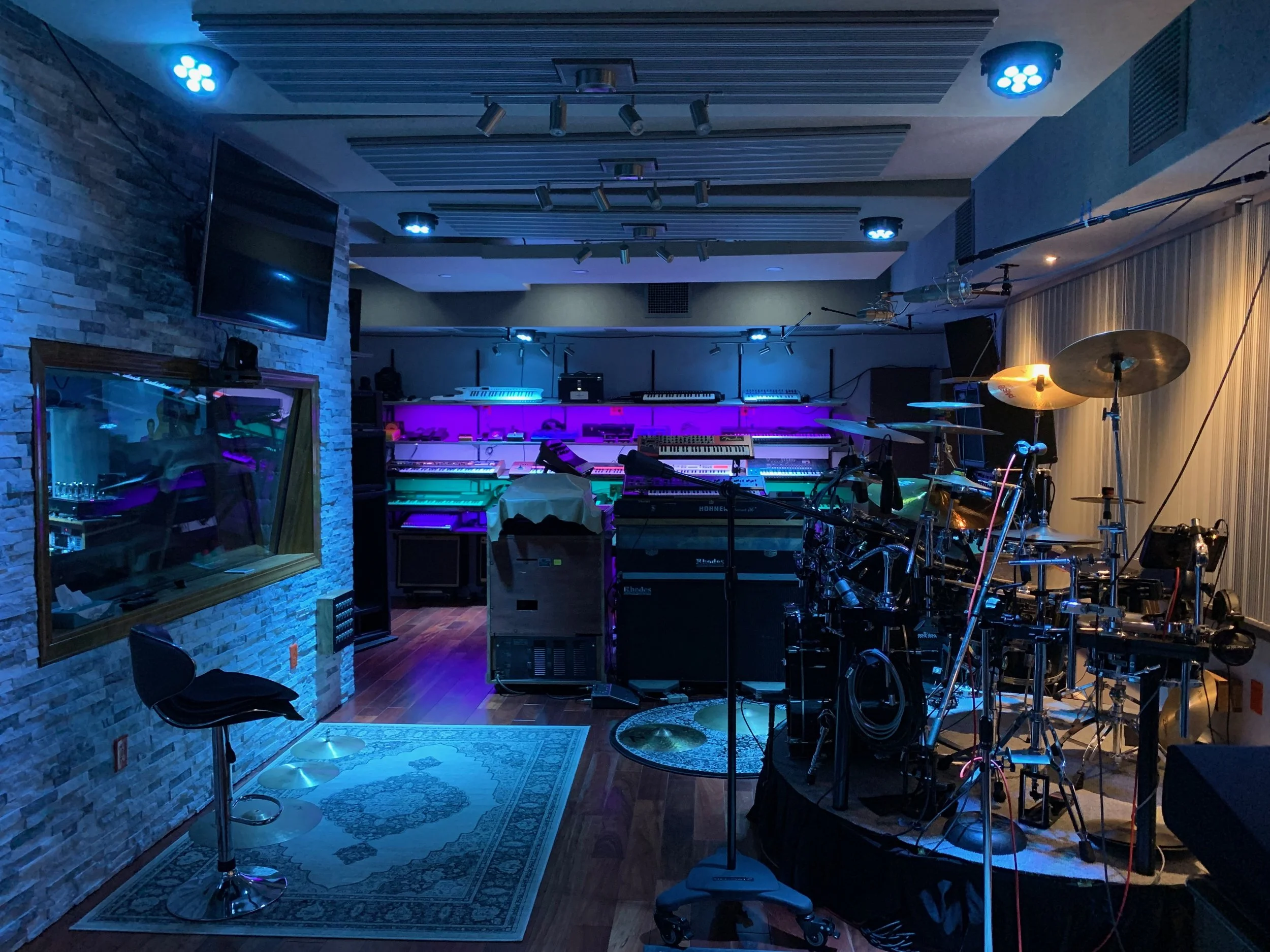 A music studio with various instruments and equipment. It features a drum set, keyboards, and microphones. The room is decorated with blue lighting and has a stone wall on one side and a patterned rug on the floor.