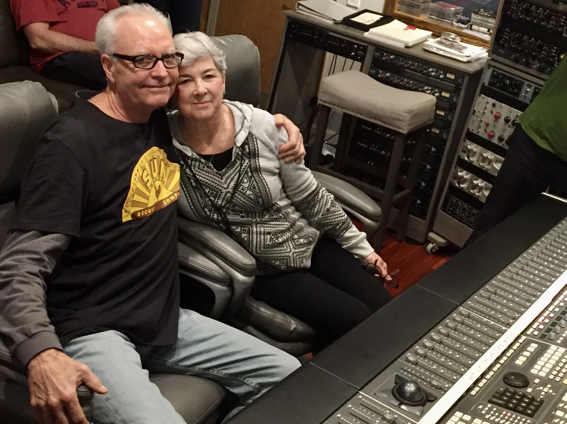 A man and a woman sitting close together in a recording studio, surrounded by audio equipment, with a mixing console in front of them.